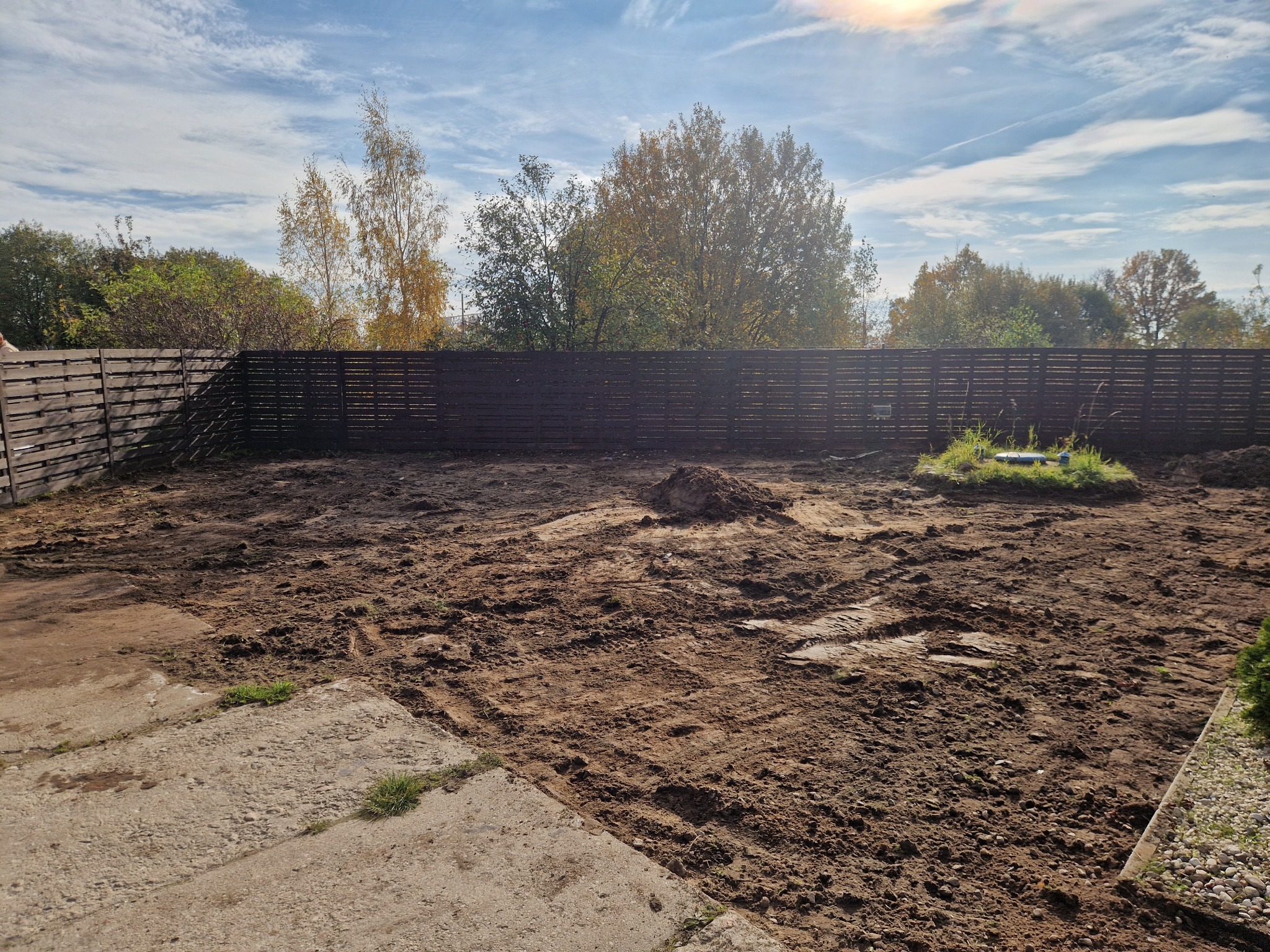 Przygotowany teren pod budowę z widocznymi śladami maszyn, ogrodzony drewnianym płotem. W tle drzewa i błękitne niebo z delikatnymi chmurami.