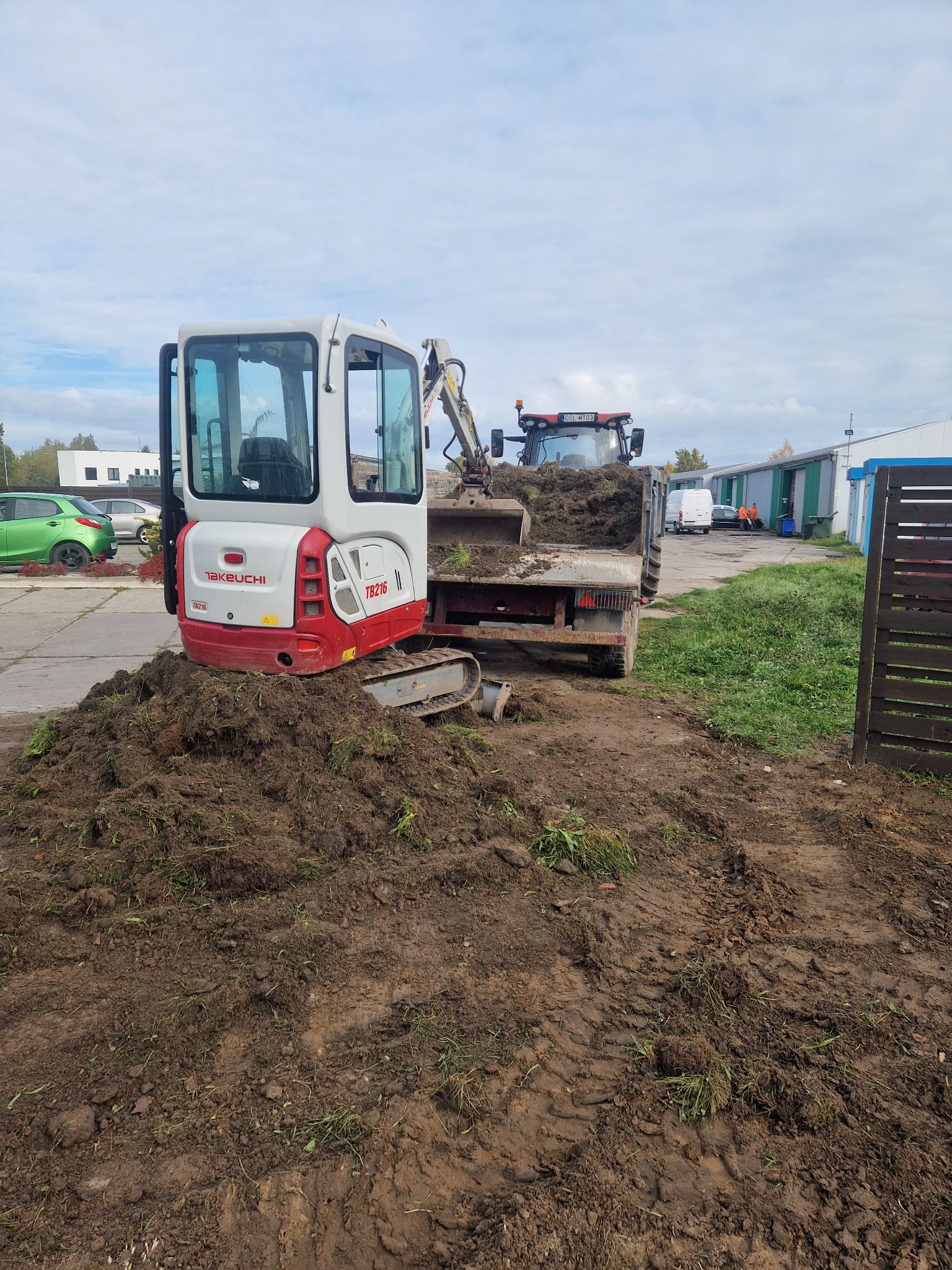 Mini koparka Takeuchi TB216 ładuje ziemię do traktora na tle zabudowań gospodarczych. Widoczny świeży nasyp ziemi.