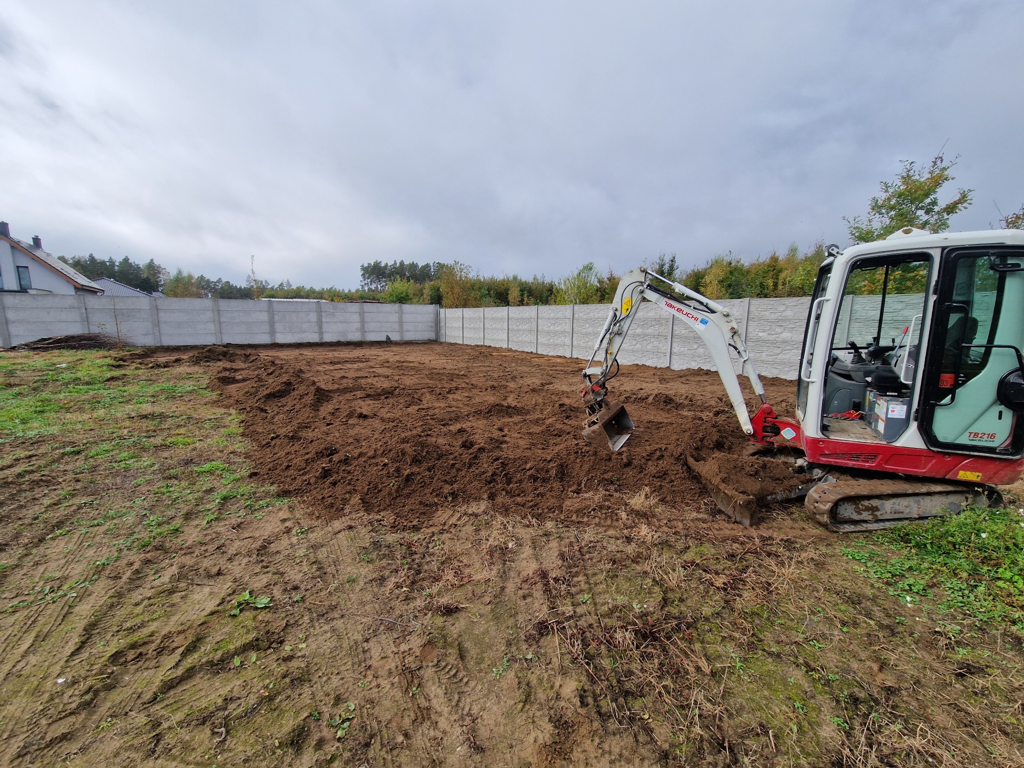 Mini koparka Takeuchi TB216 w trakcie niwelacji terenu pod budowę, widoczny wyrównany obszar ziemi i betonowe ogrodzenie w tle.