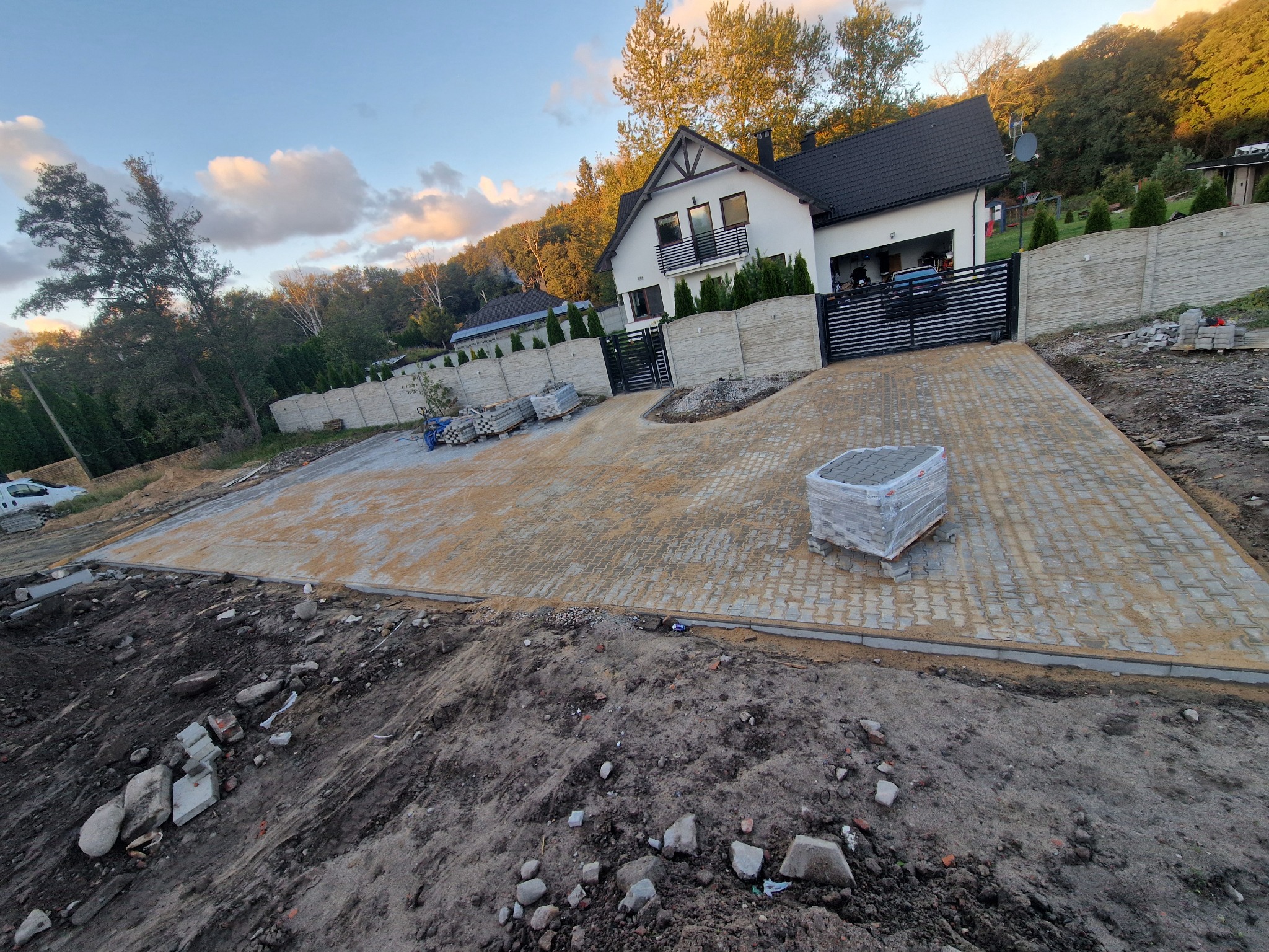 Fragment posesji z częściowo ułożoną kostką brukową w odcieniach beżu. Na pierwszym planie ziemia, w tle dom z czarnym dachem i ogrodzenie z betonu architektonicznego.