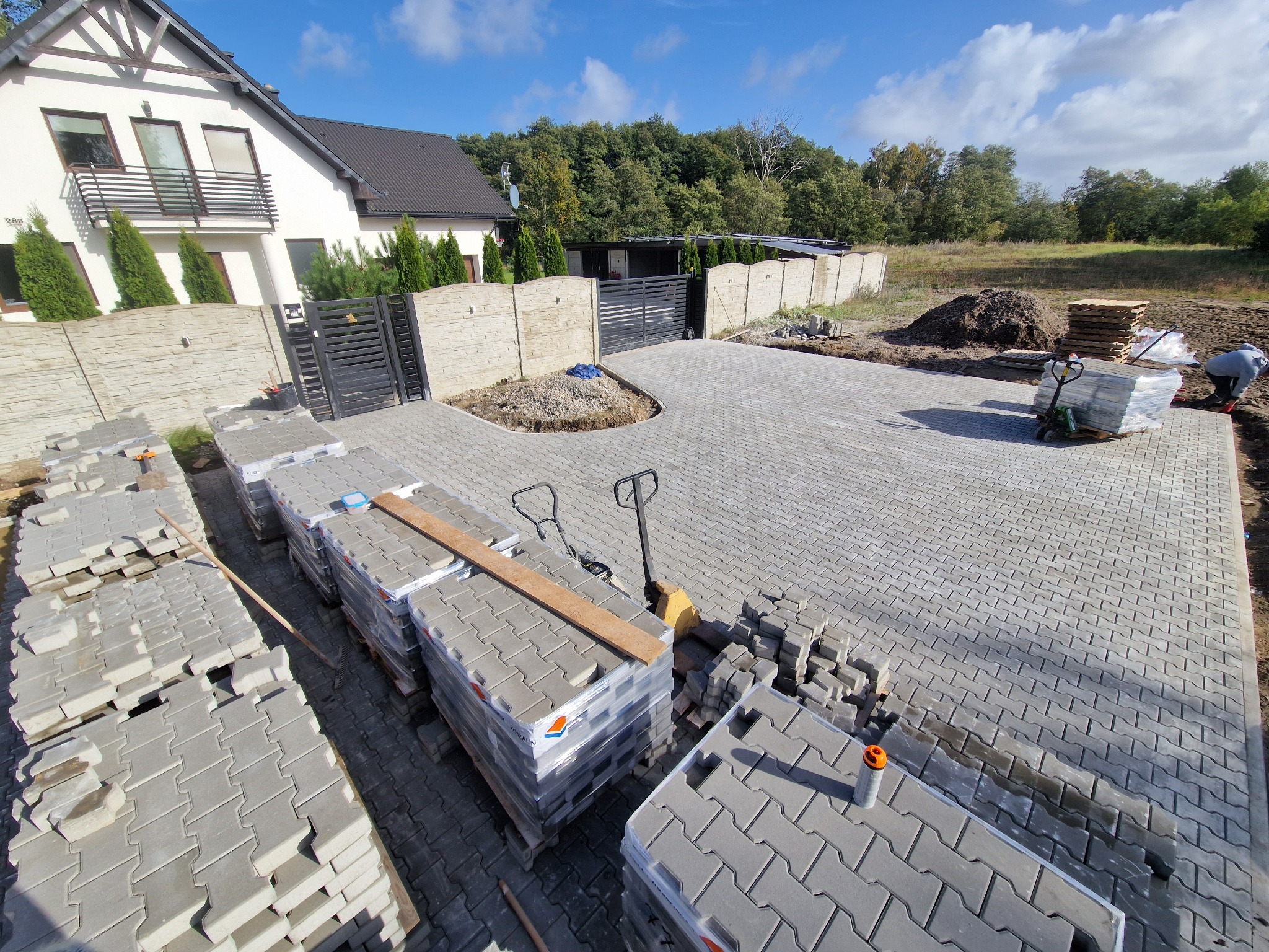Ułożona szara kostka brukowa na podjeździe domu jednorodzinnego. Palety z kostką i pracownik w tle. Widok z góry, słoneczny dzień.
