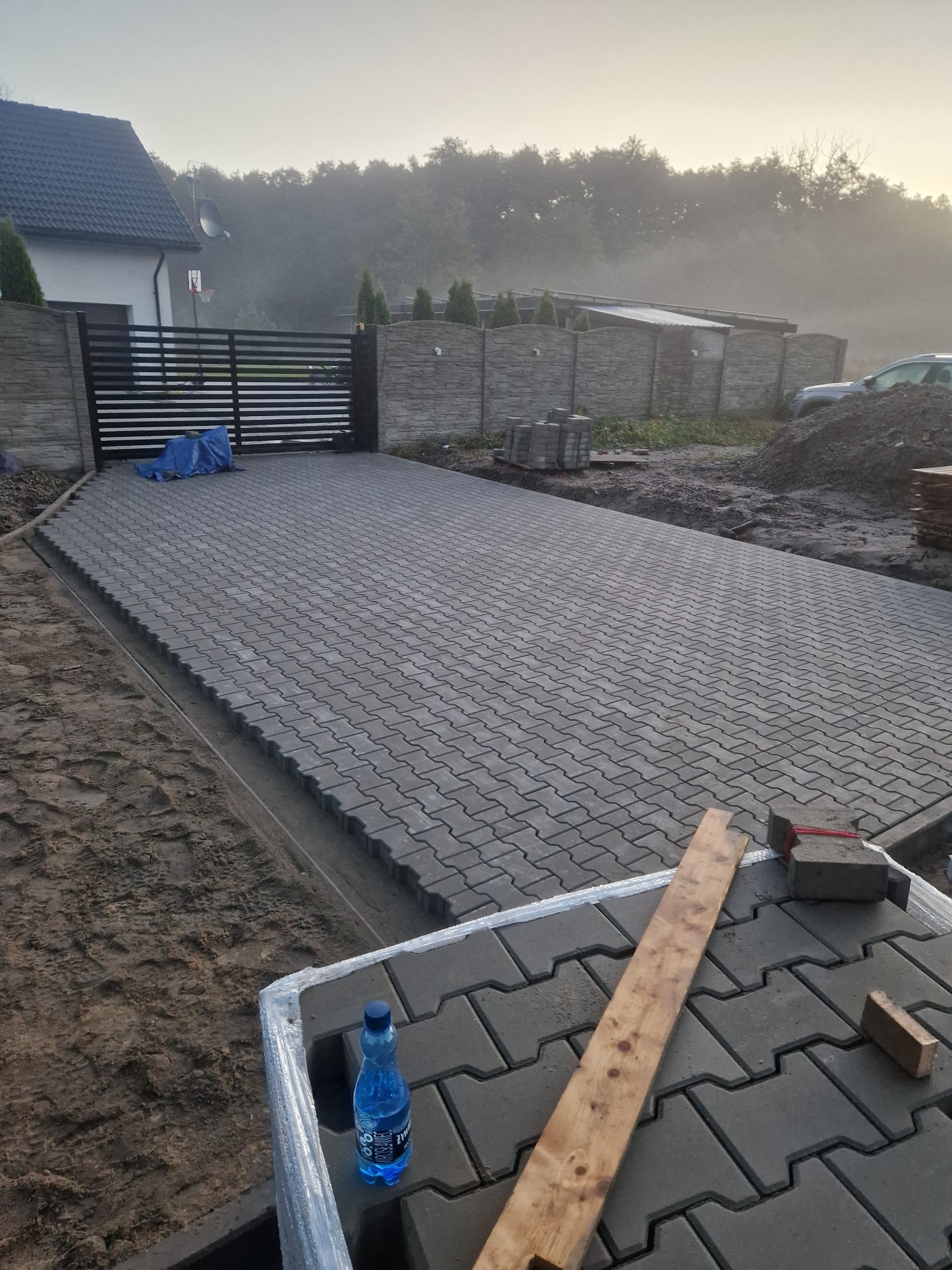 Ułożona szara kostka brukowa na podjeździe, widok z góry, w tle brama wjazdowa i dom. Na pierwszym planie paleta z kostką i butelka wody.