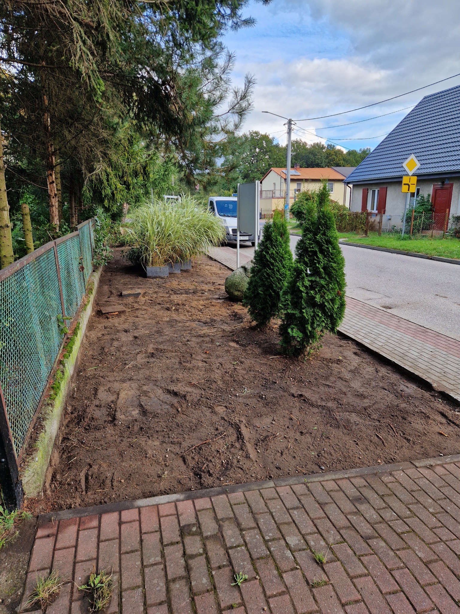 Przygotowany teren pod nasadzenia z widocznymi śladami prac ziemnych, obok chodnik z kostki, w tle budynek mieszkalny i zaparkowany biały samochód.