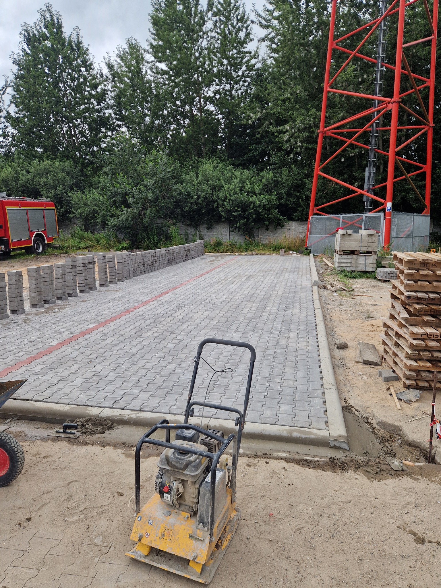 Szara kostka brukowa ułożona na parkingu, z żółtą zagęszczarką wibracyjną na pierwszym planie. W tle czerwona wieża telekomunikacyjna i wóz strażacki.