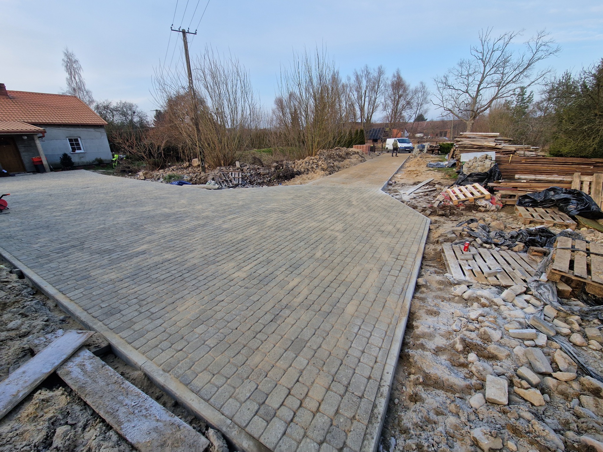 Ułożona szara kostka brukowa na podjeździe w kształcie litery Y, z widocznymi paletami i materiałami budowlanymi w tle. Częściowo ukończony projekt.