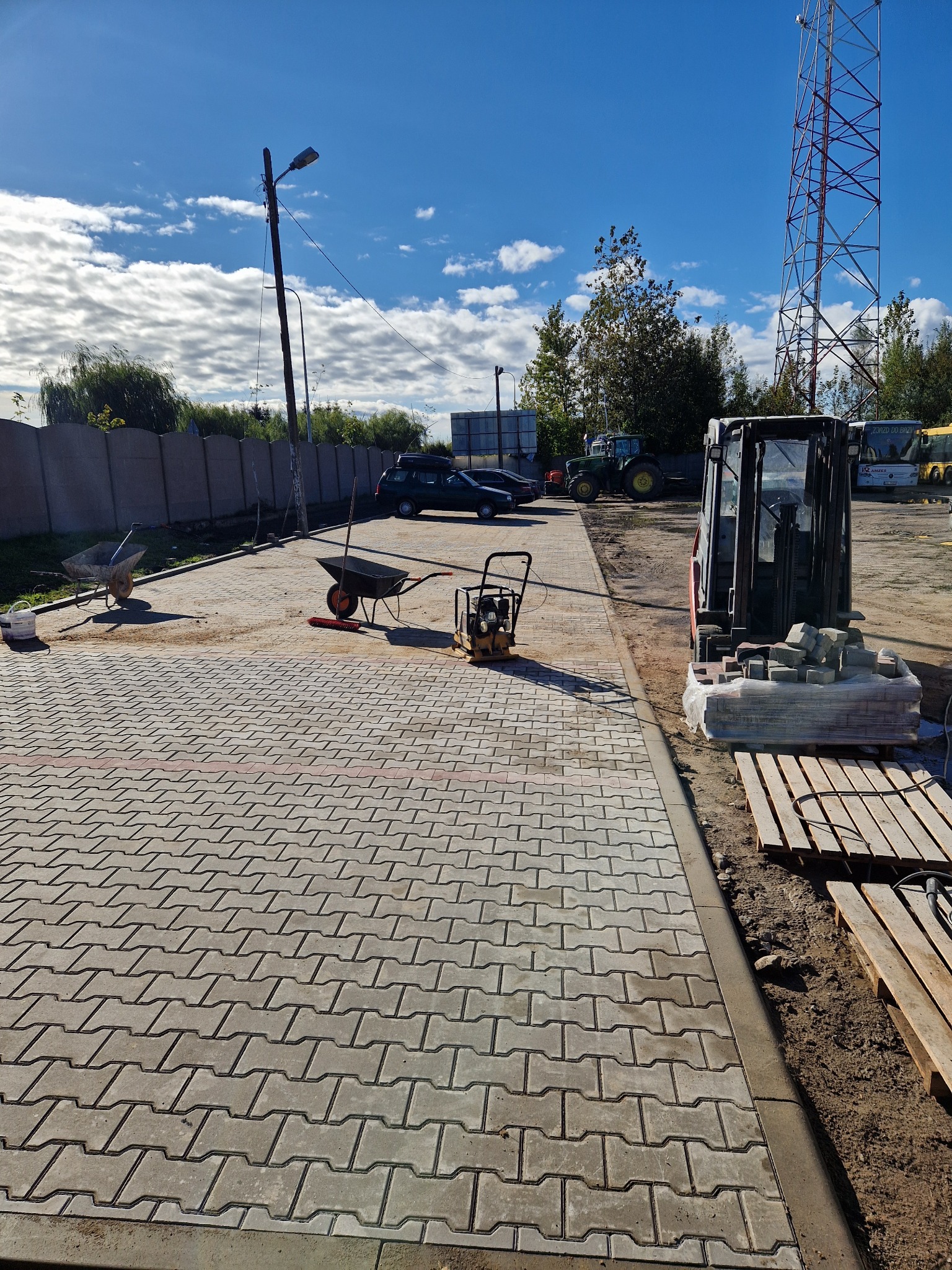 Ułożona kostka brukowa w jodełkę, z widocznymi narzędziami: wózek, ubijarka. W tle zaparkowane pojazdy i wysoka wieża telekomunikacyjna. Słoneczny dzień.