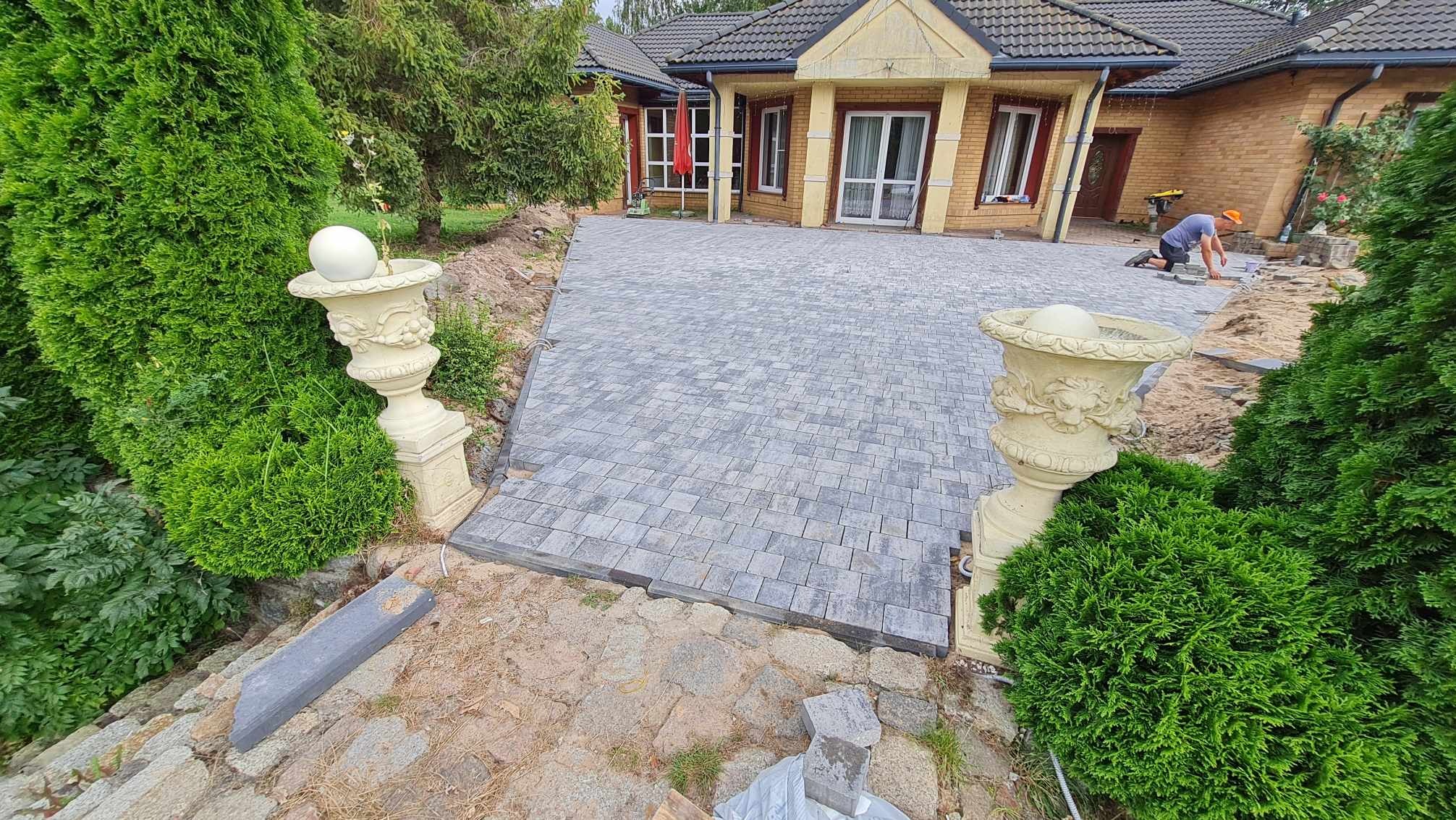Ułożona kostka brukowa w odcieniach szarości prowadząca do domu z cegły, z dekoracyjnymi wazonami po bokach i pracującym brukarzem w tle. Widok z góry.