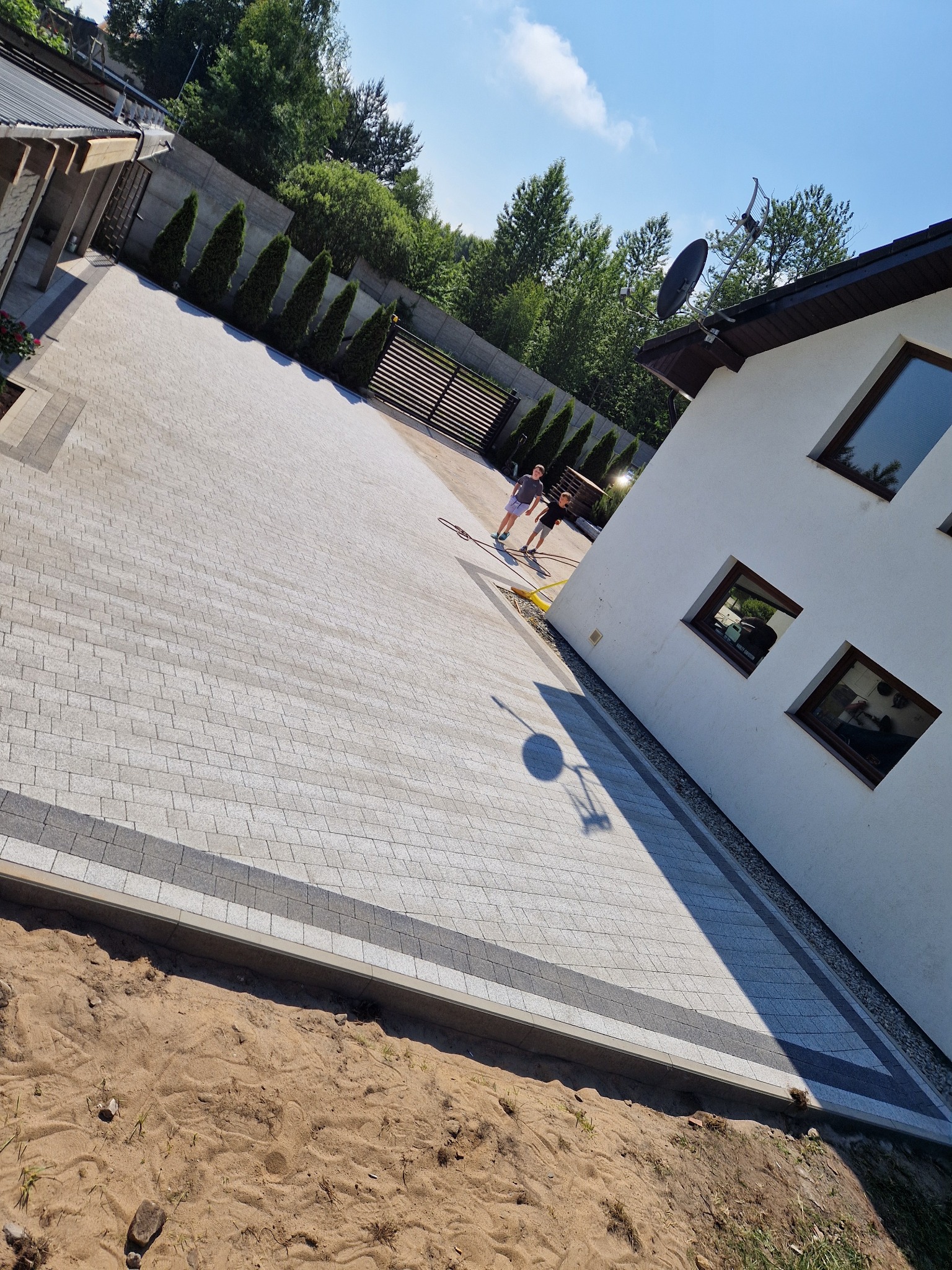 Ułożona kostka brukowa w odcieniach szarości tworzy wzór na podjeździe. Widoczne obrzeża, fragment domu z oknami i zieleń w tle. Dwóch chłopców w oddali.