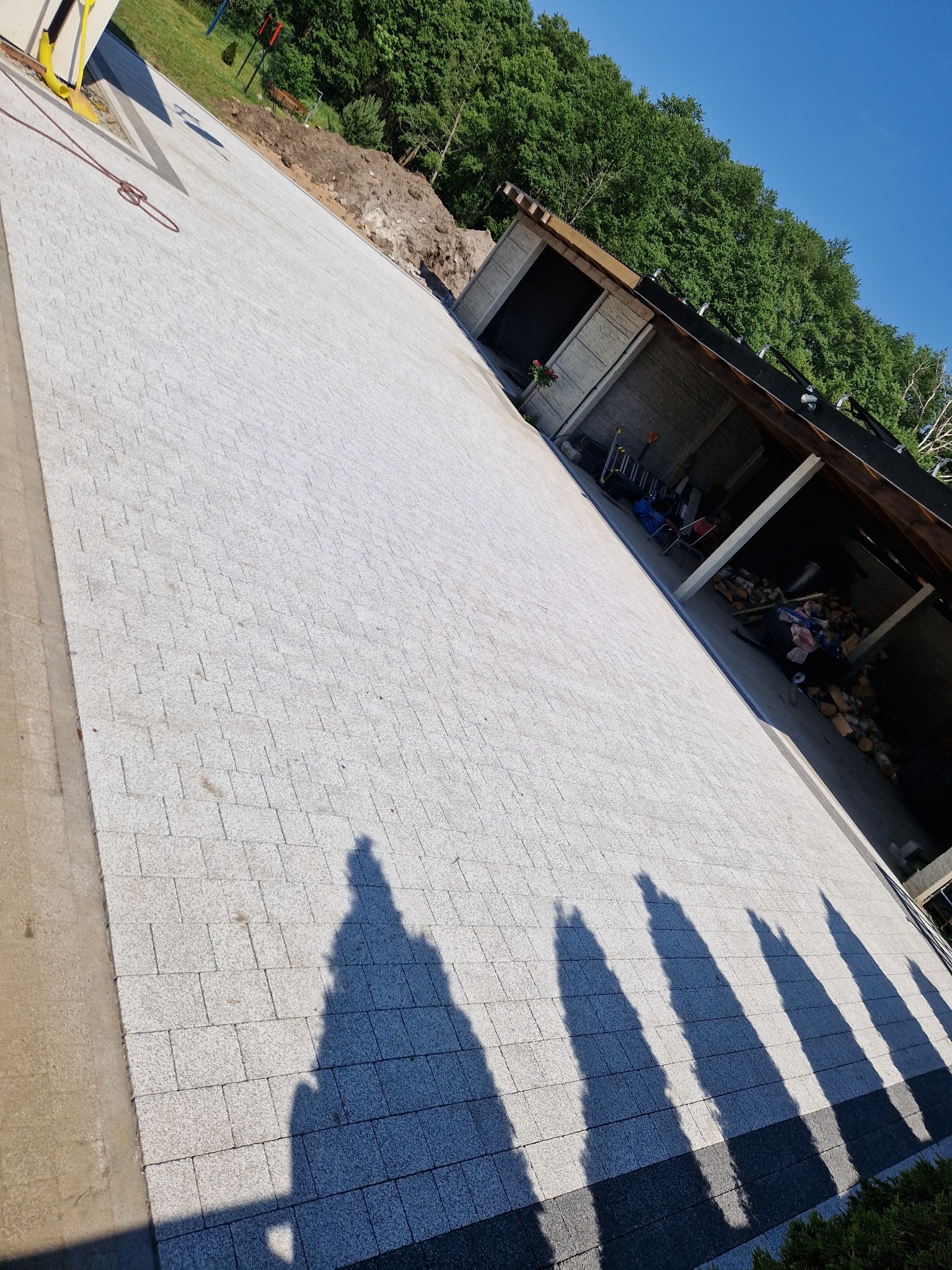 Jasnoszara kostka brukowa ułożona na posesji, z widocznymi cieniami drzew i fragmentem budynku gospodarczego w tle, w słoneczny dzień.