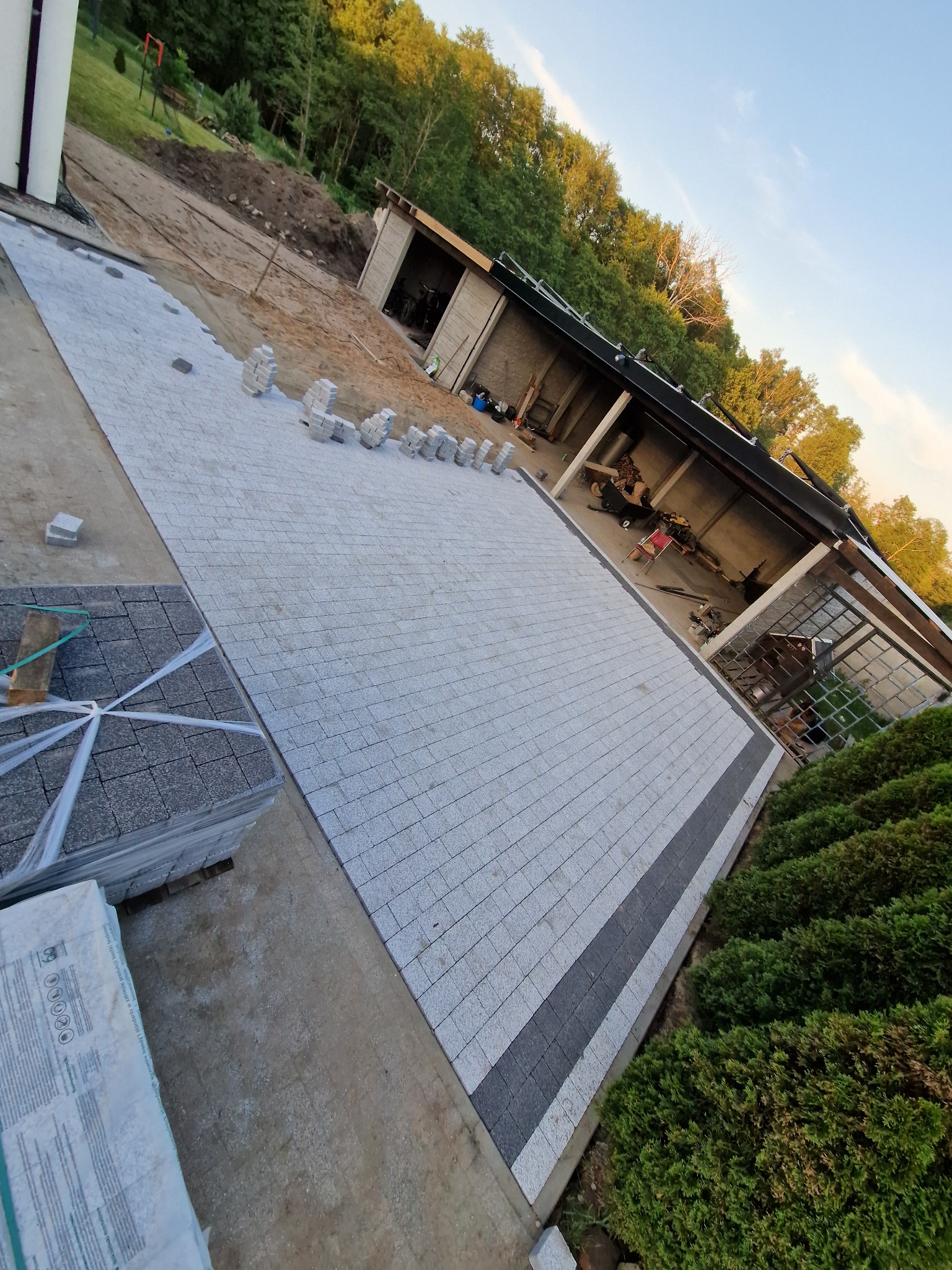 Fragment układanej szarej kostki brukowej z ciemnym pasem, obok ułożone kostki i zapas kostki w opakowaniu, w tle budynek gospodarczy i zieleń.