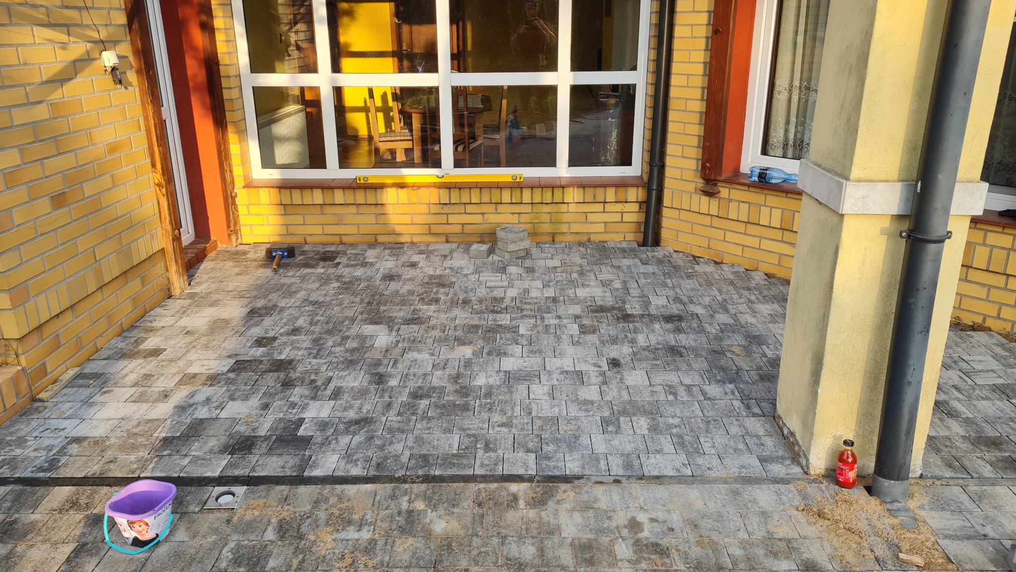 Ułożona kostka brukowa na tarasie domu z żółtej cegły. Widoczny fioletowy wiaderko z motywem z bajki Kraina Lodu i butelka soku. Częściowo widoczne okno i kolumna.