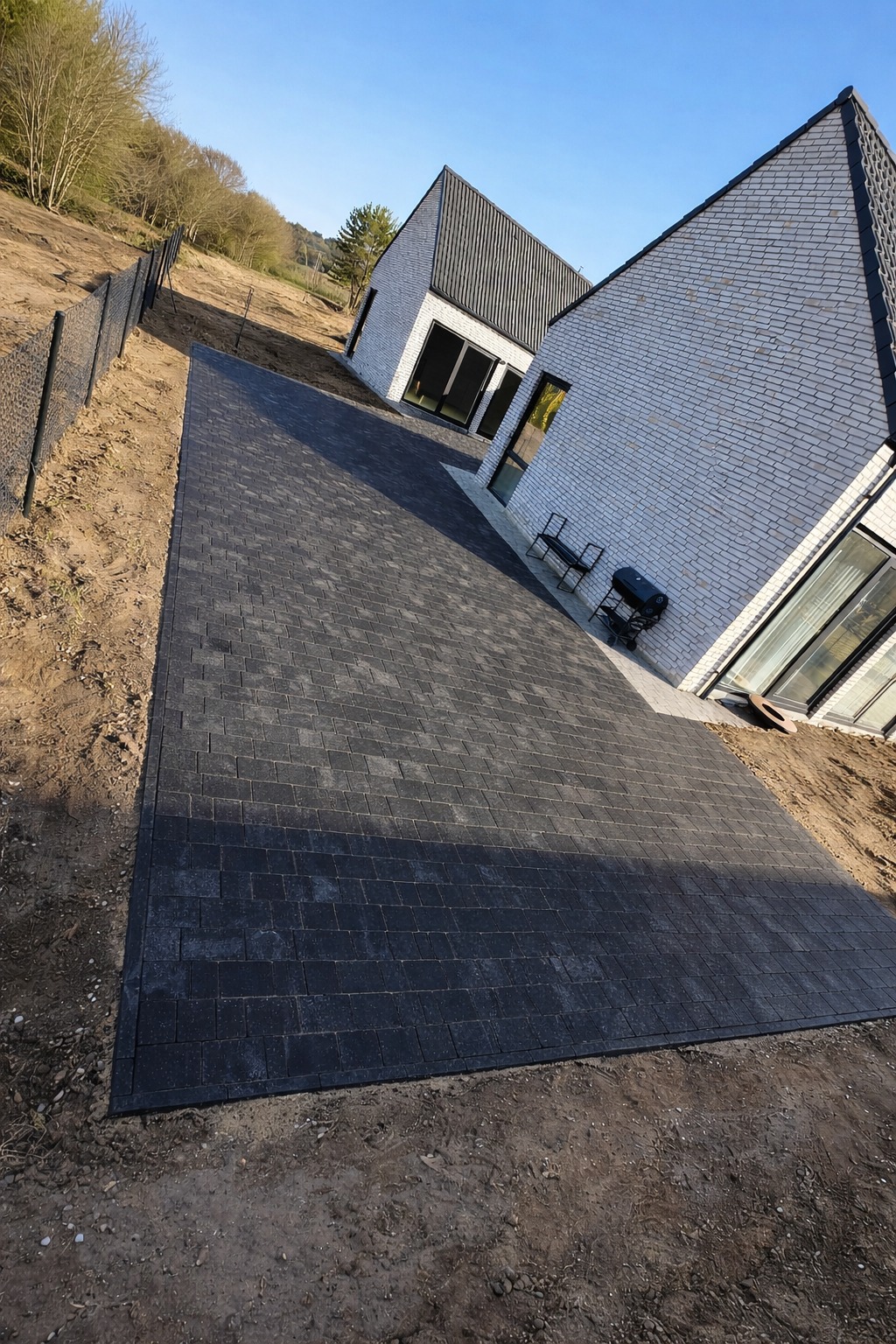 Ciemnoszara kostka brukowa ułożona na podjeździe do nowoczesnego domu z białej cegły, widok z góry, błękitne niebo w tle. Minimalistyczny design.