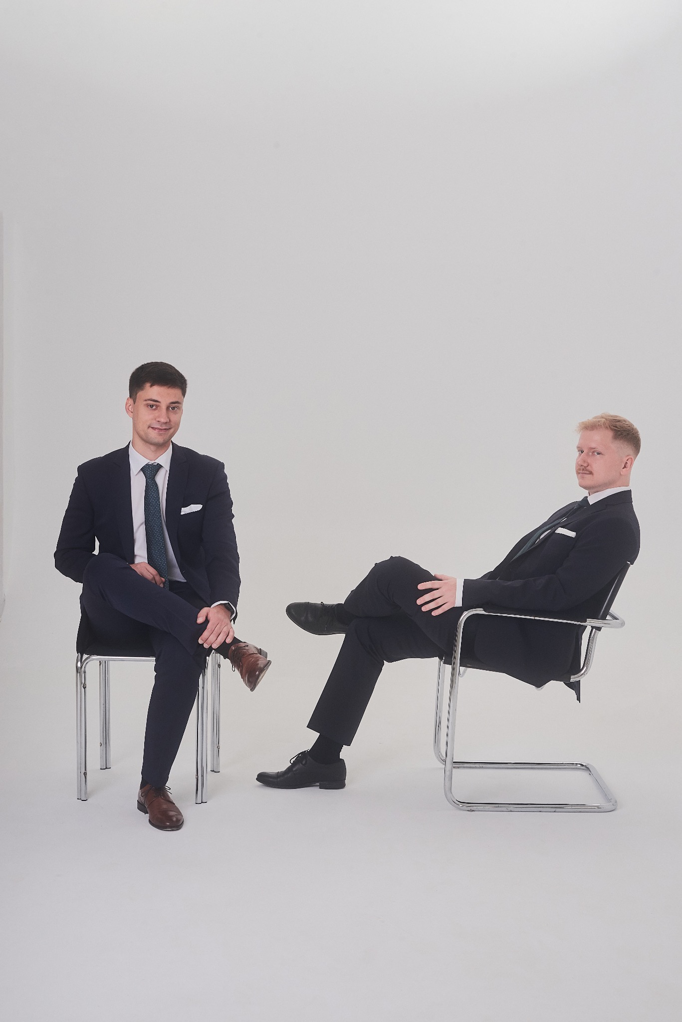 Dwóch eleganckich mężczyzn w garniturach siedzi na krzesłach na jasnym tle. Jeden patrzy w kamerę, drugi odwrócony bokiem. Profesjonalny wizerunek.