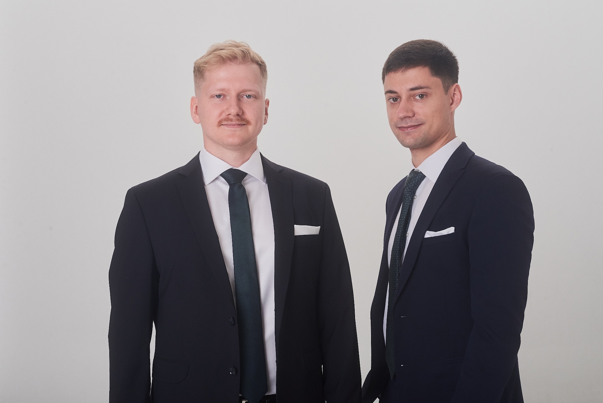 Portret dwóch elegancko ubranych mężczyzn w garniturach na jasnym tle. Jeden z nich ma wąsy i blond włosy, drugi ciemne włosy. Obaj patrzą w obiektyw.