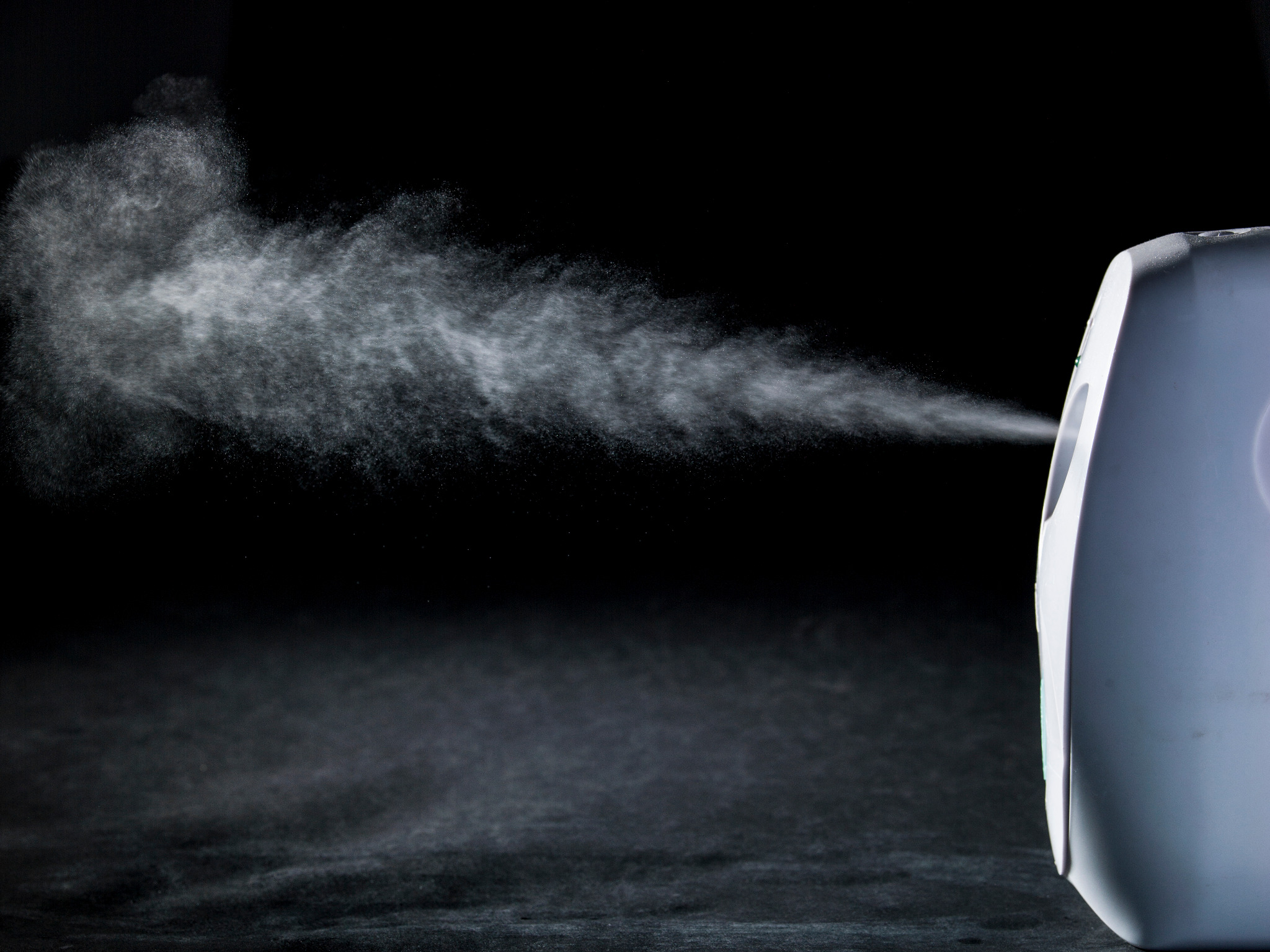 Biały dyfuzor rozpyla delikatną mgiełkę na ciemnym tle, tworząc efekt wizualny. Urządzenie ustawione z prawej strony kadru.