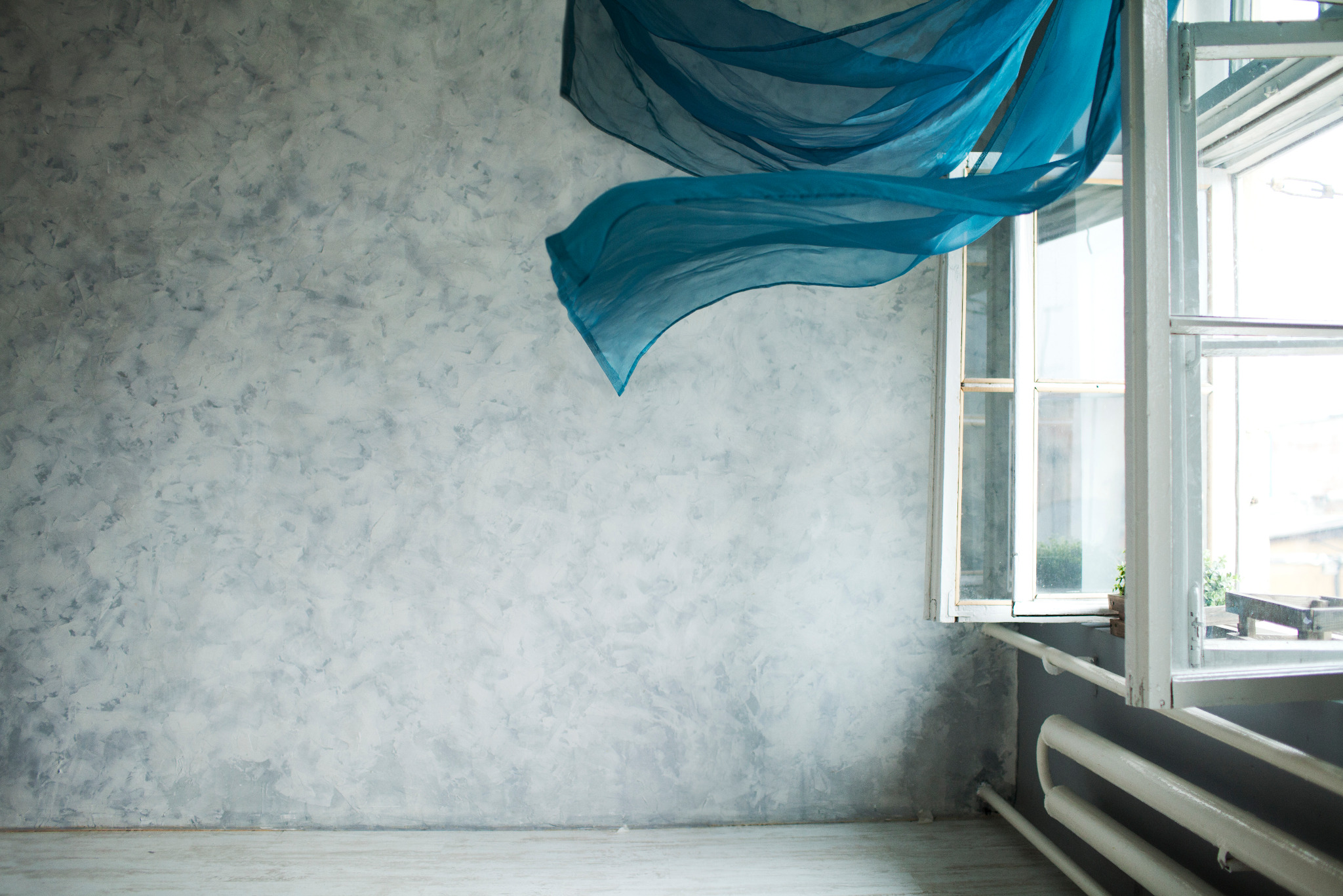 Jasne wnętrze z teksturowaną szarą ścianą, otwartym oknem i powiewającą turkusową tkaniną. Widoczny grzejnik pod oknem. Minimalistyczny wystrój.