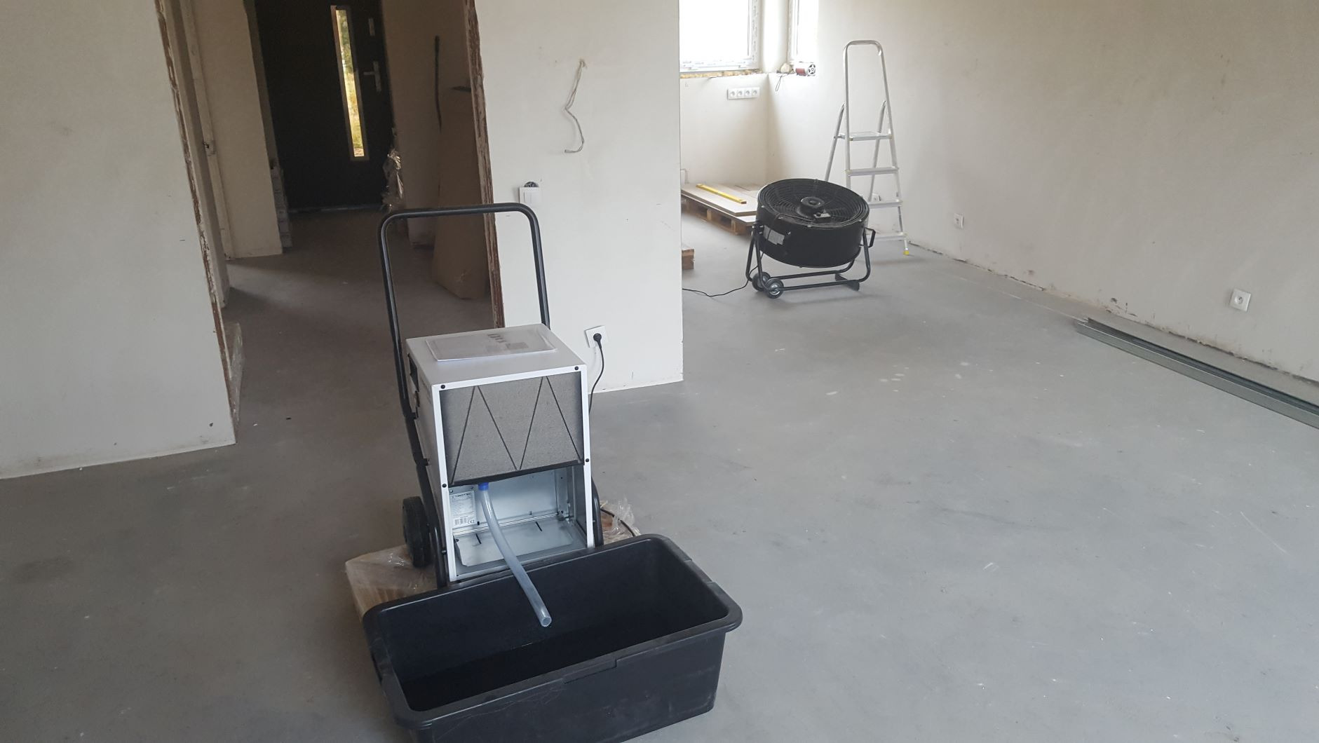 Przenośny kondensacyjny sprzęt z pojemnikiem na wodę w surowym wnętrzu. W tle wentylator przemysłowy i drabina. Widoczne ślady wilgoci na ścianach.