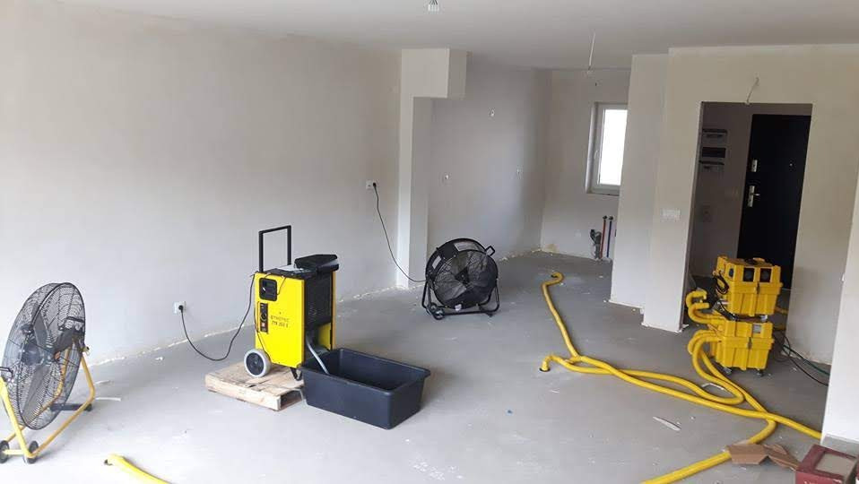 Wnętrze w trakcie prac. Żółte urządzenia i wentylatory wspomagają proces. Widoczne ślady wilgoci na ścianach i podłodze. Przewody i akcesoria rozłożone na podłodze.