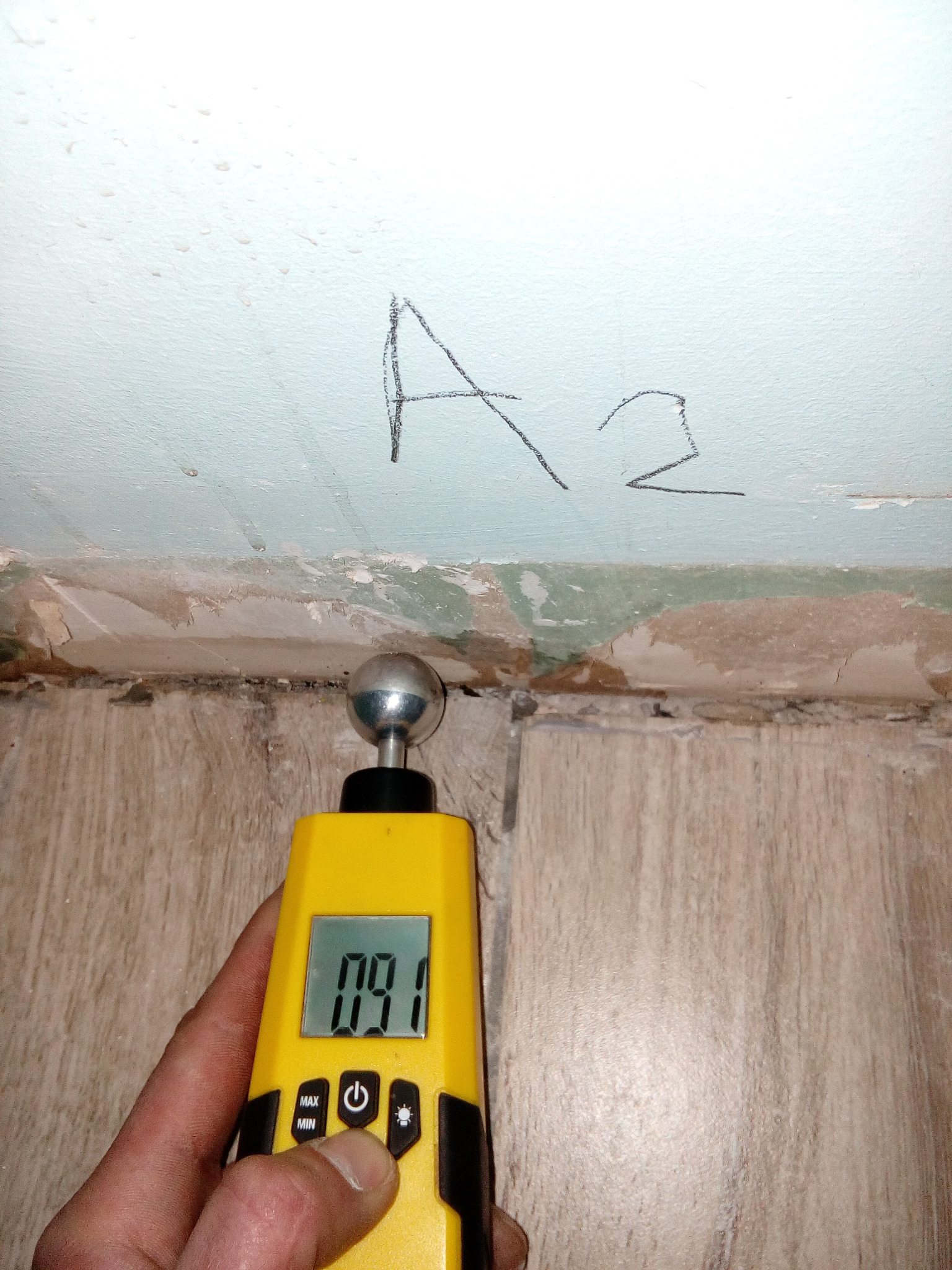 Pomiar wilgotności ściany przy podłodze za pomocą wilgotnościomierza elektronicznego. Na ścianie widoczny napis 'A2'.