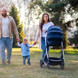 Patryk Kołodziejczyk - Uśmiechnięta rodzina spaceruje po trawniku w parku. Tata w okularach, mama z wózkiem i małe dziecko trzymające rodziców za ręce. Ciepłe, naturalne światło.