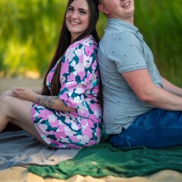 Patryk Kołodziejczyk - Uśmiechnięta para siedzi plecami do siebie na kocu na piasku, kobieta w kwiecistej sukience, mężczyzna w niebieskiej koszuli, w tle zieleń. Sesja plenerowa.