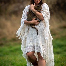 Patryk Kołodziejczyk - Kobieta w białej sukience i wianku na głowie, w okularach, trzymająca kota na tle zielonej trawy i brązowego tła. Ujęcie w stylu sesji kobiecej.