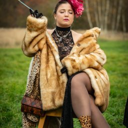 Patryk Kołodziejczyk - Kobieta w futrze i kapeluszu z piórami siedzi na krześle na łące, trzymając lufkę. Ubrana w koronkową bluzkę i buty w panterkę. Stylizacja retro, sesja plenerowa.