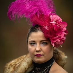 Patryk Kołodziejczyk - Portret kobiety w czarnej koronkowej sukience, futrzanym szalu i kapeluszu z różowych piór i kwiatów na rozmytym tle. Wyraziste makijaż i biżuteria.
