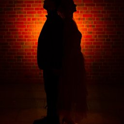 Patryk Kołodziejczyk - Sylwetki pary na tle ceglanej ściany oświetlonej ciepłym światłem. Mężczyzna w ciemnym ubraniu, kobieta w sukni. Ujęcie w ciemnej tonacji, podkreślające kontury postaci.