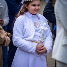 Patryk Kołodziejczyk - Dziewczynka w białej albie komunijnej, z wiankiem na głowie i świecą w dłoniach, uśmiecha się delikatnie. W tle widoczni goście w strojach wizytowych.