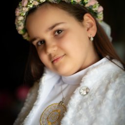 Patryk Kołodziejczyk - Portret dziewczynki w wianku z róż i białym stroju komunijnym, z delikatnym uśmiechem i zawieszką IHS na szyi, na ciemnym tle. Subtelne oświetlenie podkreśla detale.