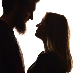 Patryk Kołodziejczyk - Silhouetted para zakochanych patrzy na siebie na jasnym tle. Mężczyzna z brodą i włosami związanymi w kok. Kobieta z długimi włosami.