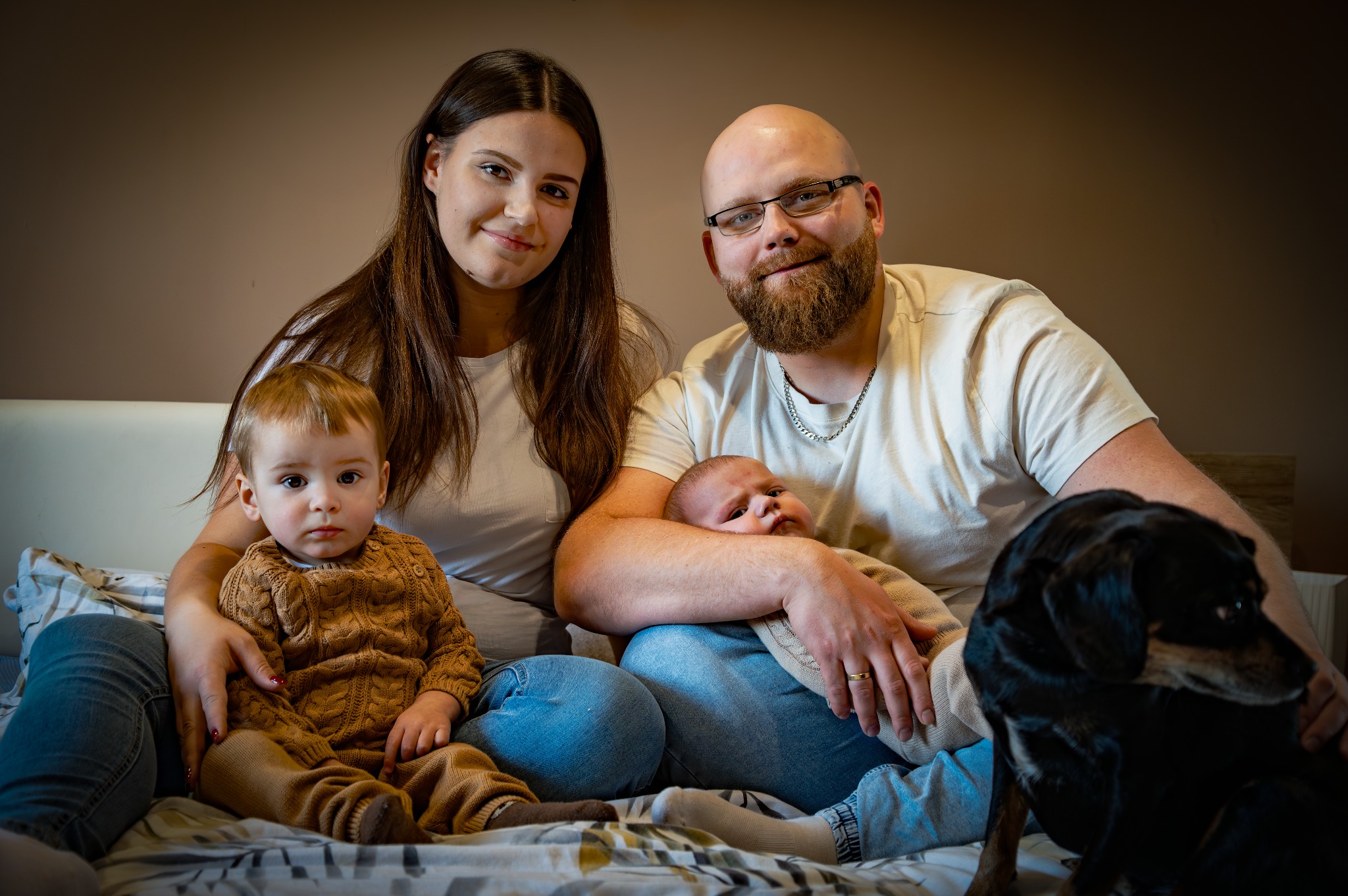 Rodzinne ujęcie: rodzice z dwójką małych dzieci i psem na łóżku. Uśmiechnięta mama, tata w okularach i z brodą, mały chłopiec w swetrze i niemowlę w ramionach taty.