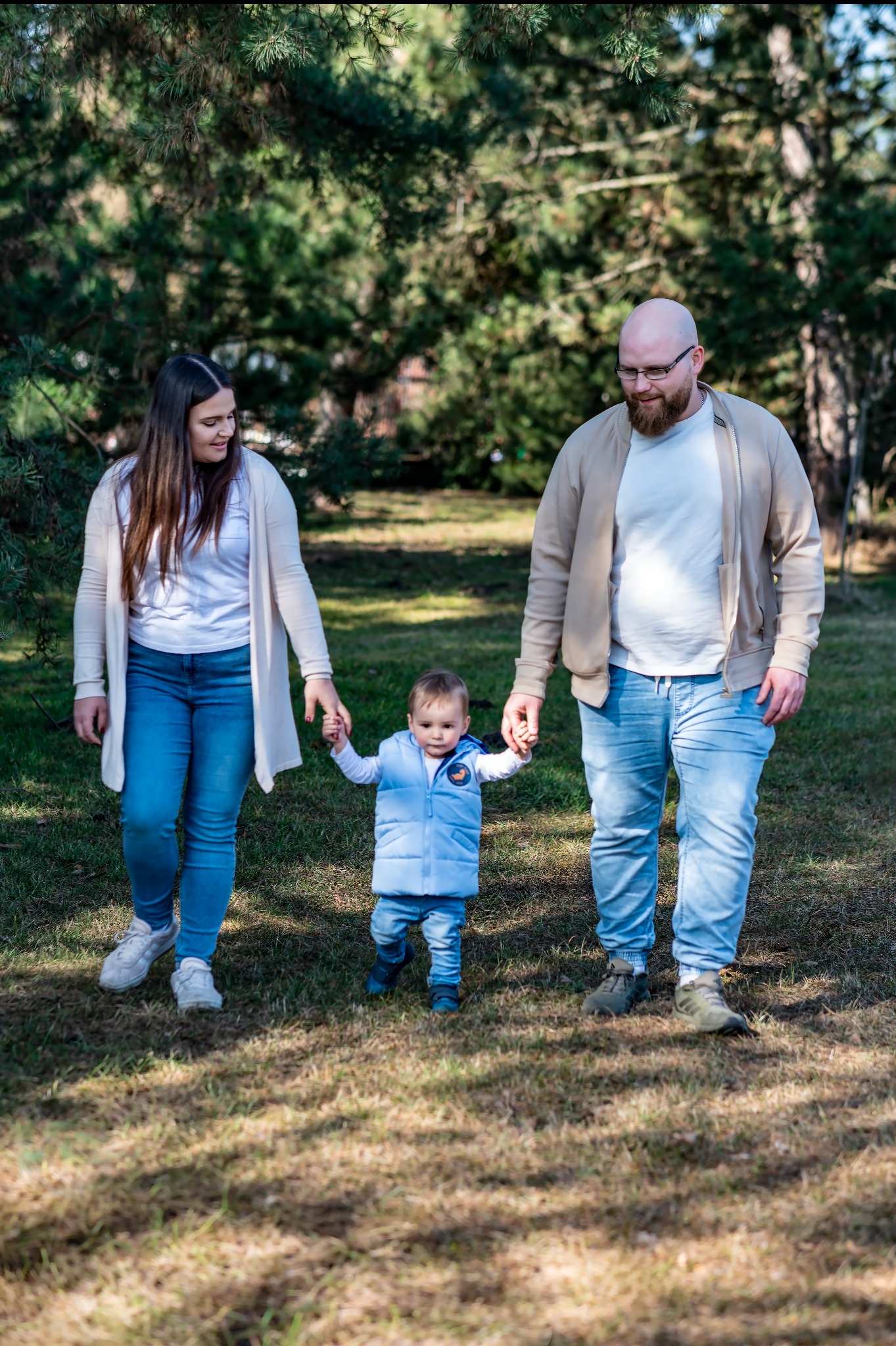 Rodzinny spacer: Mama, tata i mały syn trzymają się za ręce, idąc po trawie w słoneczny dzień. W tle drzewa iglaste. Ubrani w jasne, codzienne stroje.