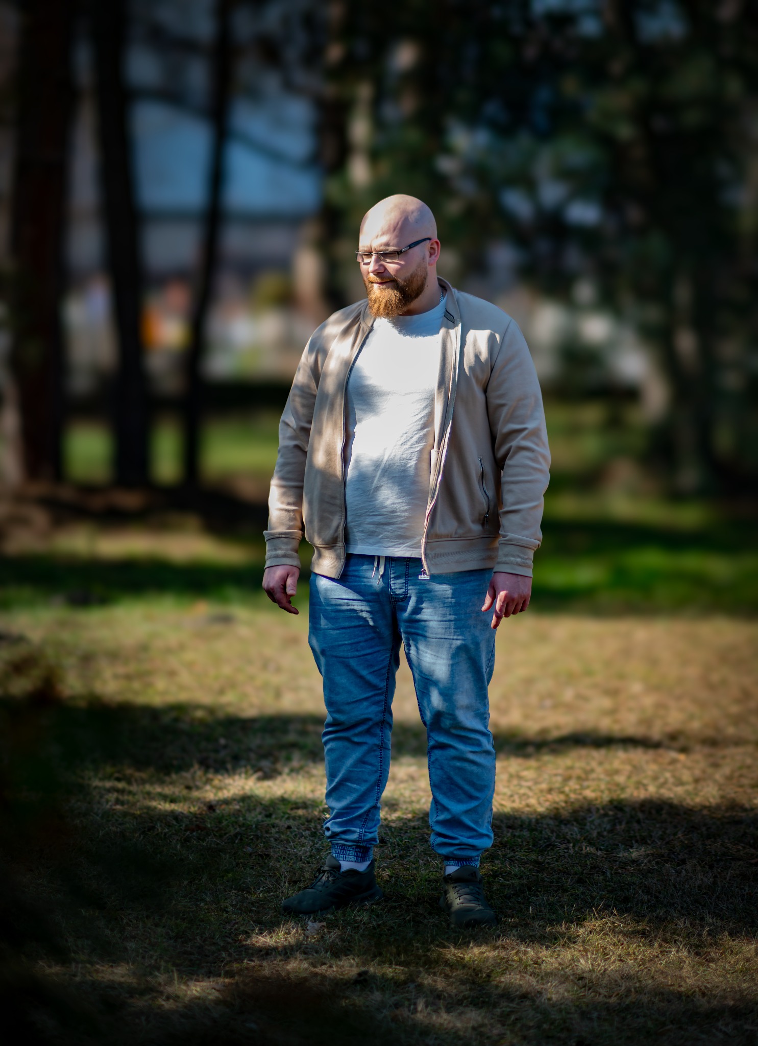 Mężczyzna z rudą brodą i okularach, ubrany w jeansy i beżową bluzę, stoi na trawie w parku w słoneczny dzień. Ujęcie z dołu, z rozmytym tłem drzew.