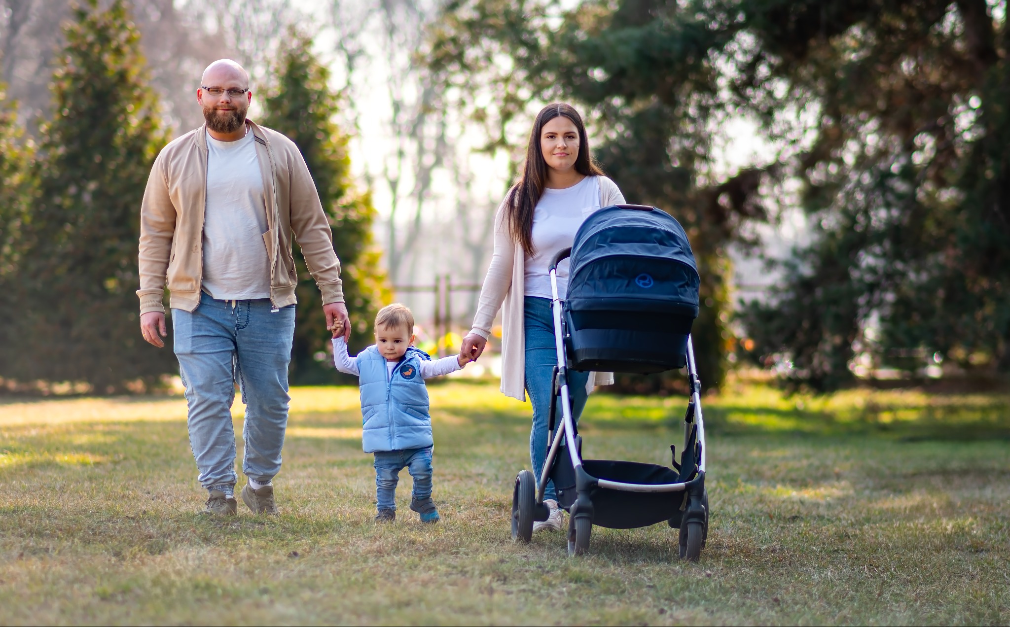 Uśmiechnięta rodzina spaceruje po trawniku w parku. Tata w okularach, mama z wózkiem i małe dziecko trzymające rodziców za ręce. Ciepłe, naturalne światło.