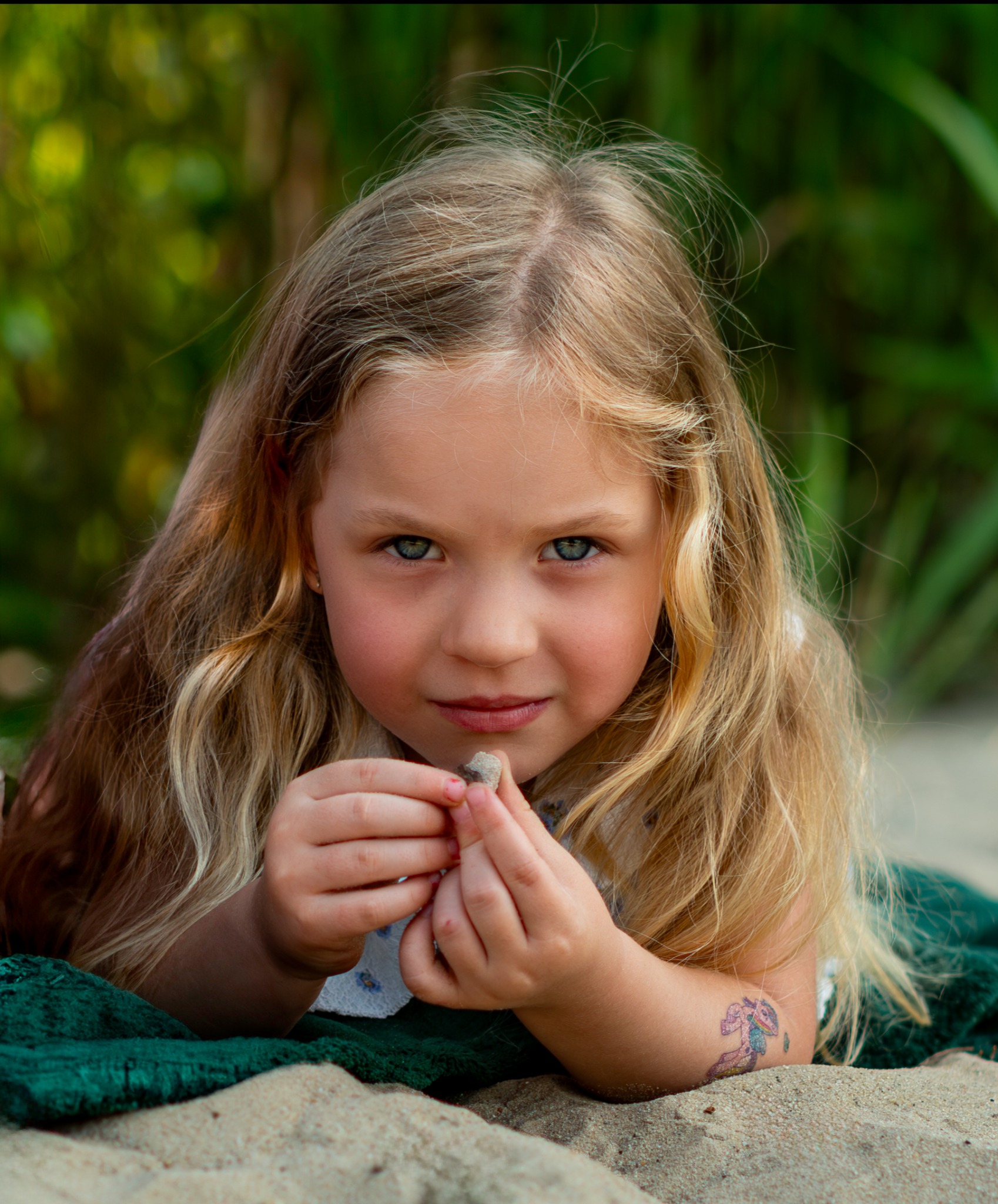 Portret uroczej dziewczynki z blond włosami, leżącej na piasku z zielonym kocem w tle, trzymającej mały kamień w dłoniach. Widoczny tatuaż na ramieniu.