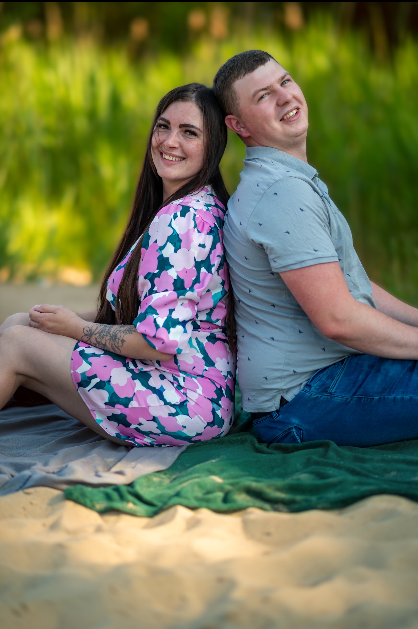 Uśmiechnięta para siedzi plecami do siebie na kocu na piasku, kobieta w kwiecistej sukience, mężczyzna w niebieskiej koszuli, w tle zieleń. Sesja plenerowa.