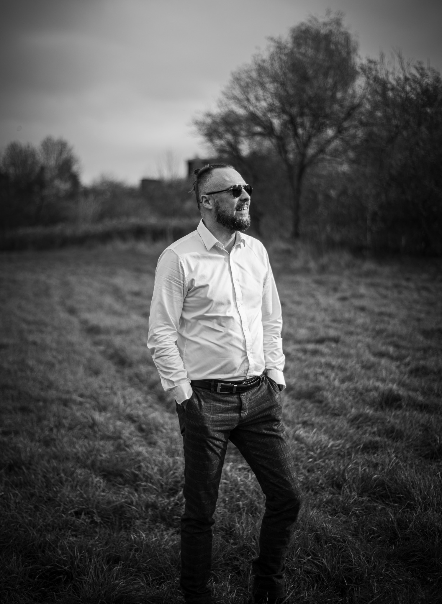 Czarno-białe zdjęcie uśmiechniętego mężczyzny w okularach przeciwsłonecznych, stojącego na łące w koszuli i spodniach w kratę, z rękami w kieszeniach.