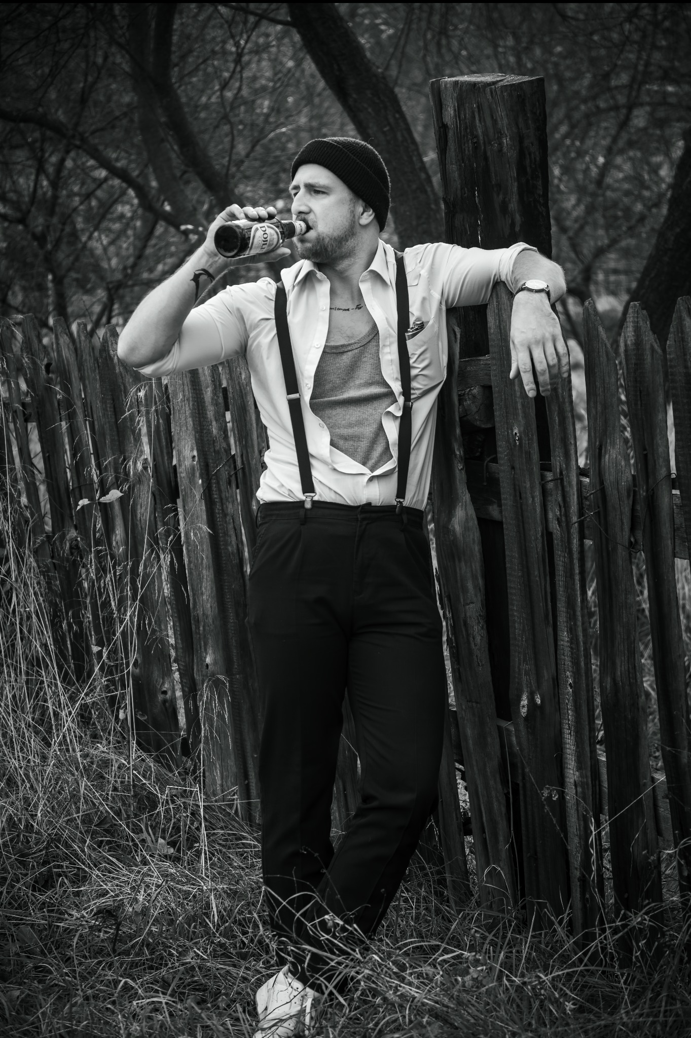 Czarno-białe zdjęcie mężczyzny w kapeluszu, pijącego piwo opierając się o drewniany płot. Ubrany w koszulę, spodnie na szelkach i sportowe buty. Tło stanowią drzewa.