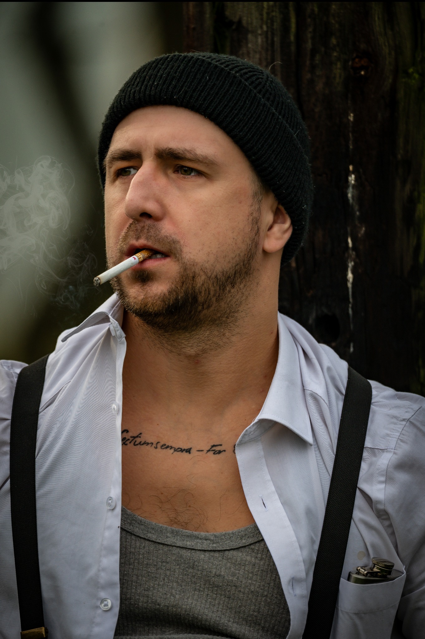 Mężczyzna w czapce i koszuli na szelkach, z papierosem w ustach, oparty o drewniany słup. Widoczny tatuaż na klatce piersiowej i piersiówka w kieszeni koszuli.