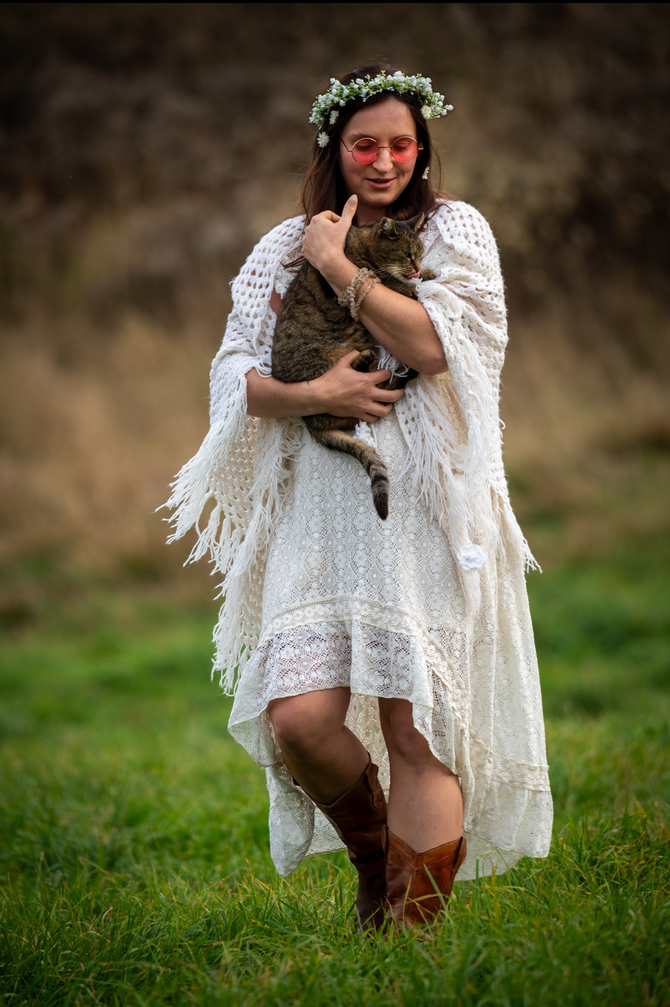 Kobieta w białej sukience i wianku na głowie, w okularach, trzymająca kota na tle zielonej trawy i brązowego tła. Ujęcie w stylu sesji kobiecej.