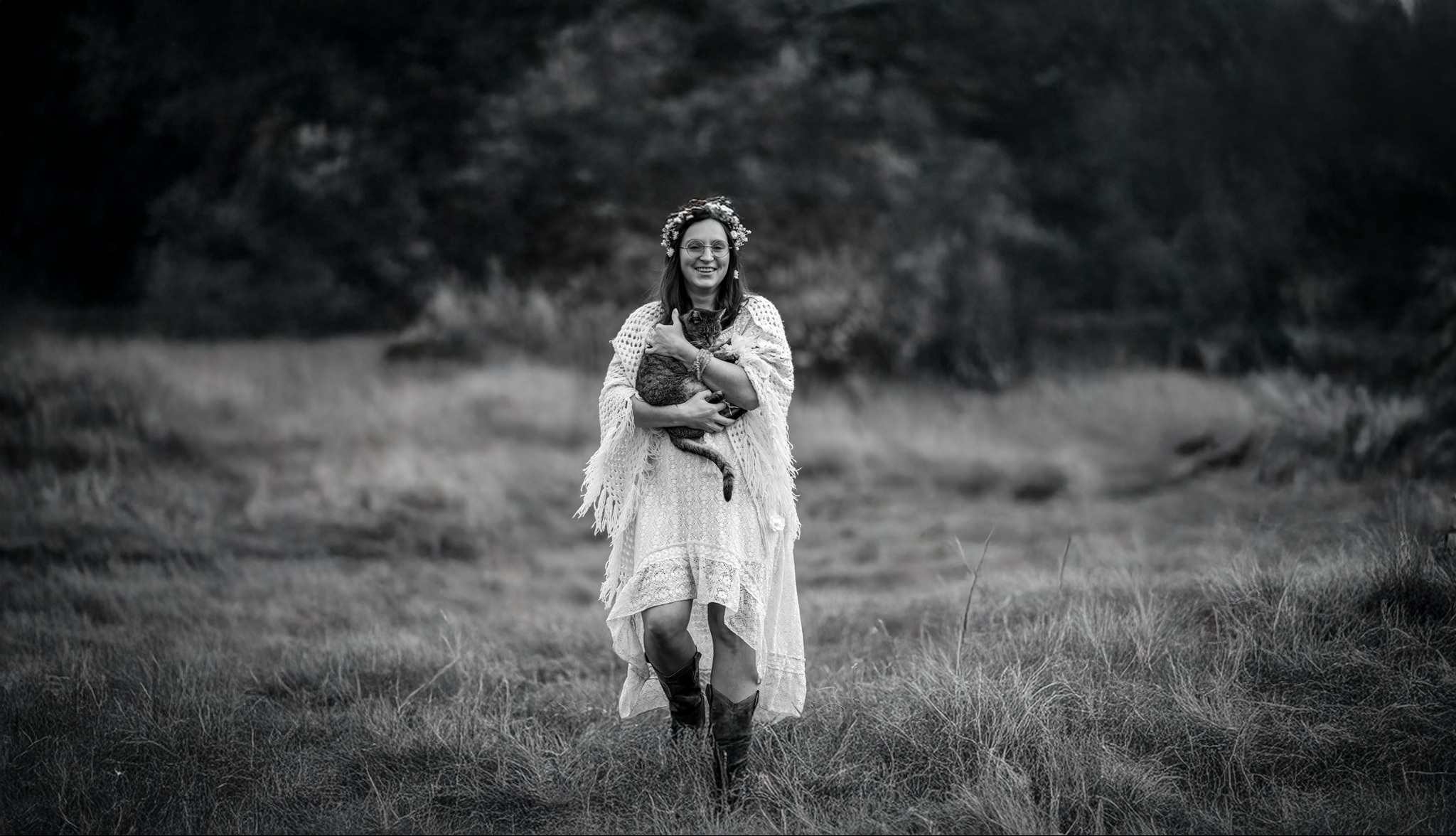 Czarno-białe zdjęcie kobiety w sukience boho i wianku, trzymającej kota na łące. Uśmiechnięta, w okularach, idzie w kierunku obiektywu. Romantyczna atmosfera.