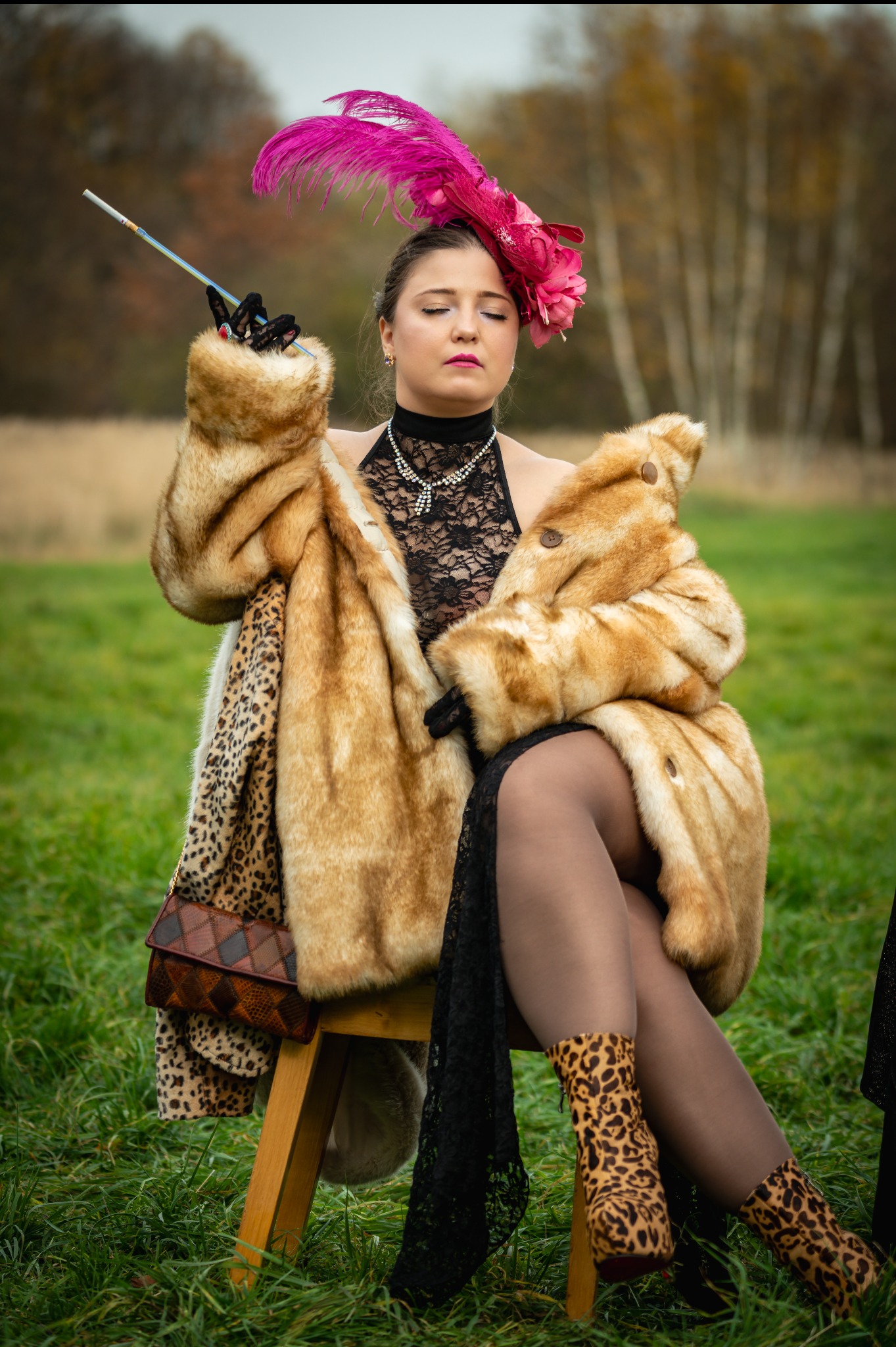 Kobieta w futrze i kapeluszu z piórami siedzi na krześle na łące, trzymając lufkę. Ubrana w koronkową bluzkę i buty w panterkę. Stylizacja retro, sesja plenerowa.