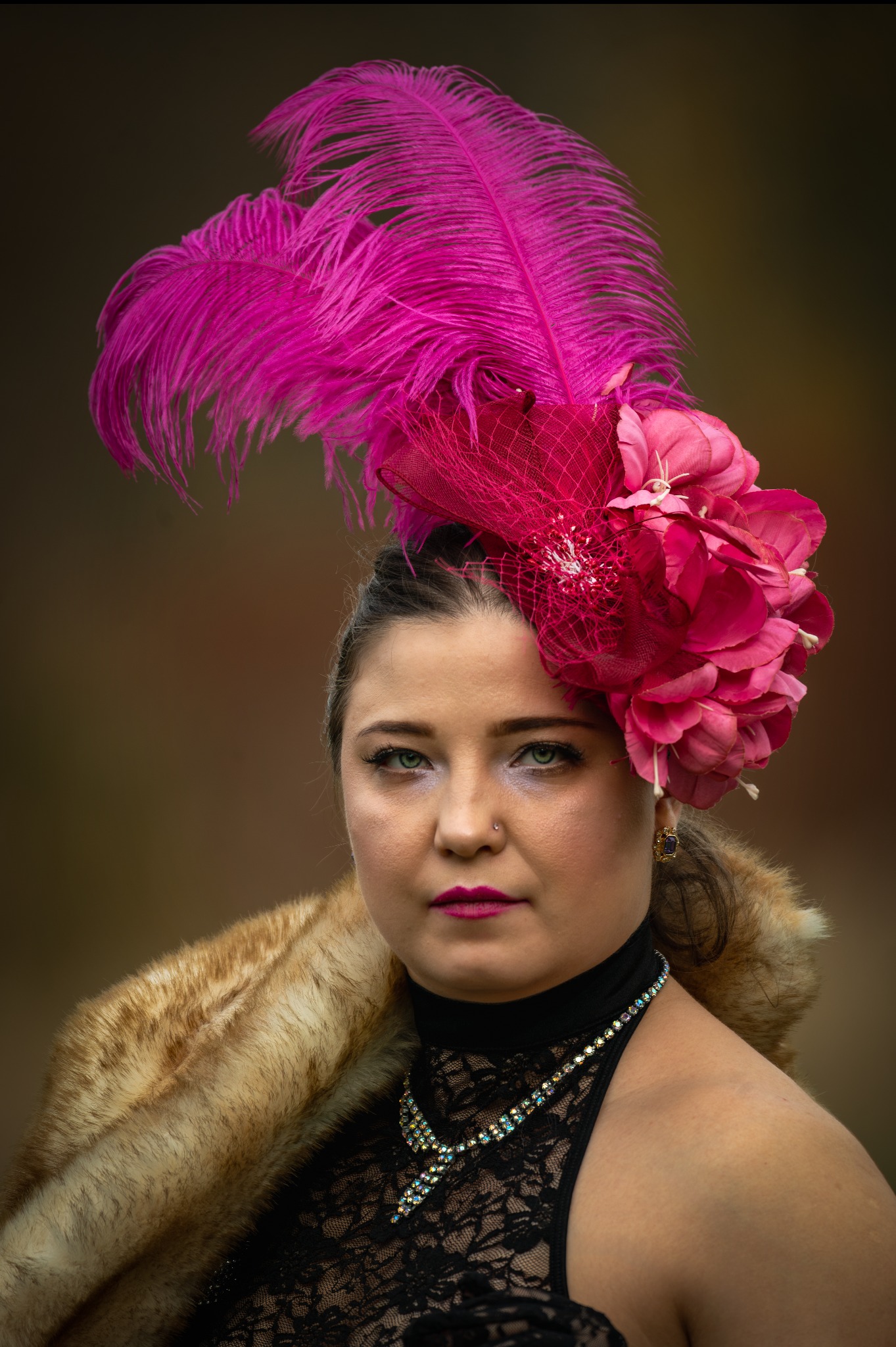 Portret kobiety w czarnej koronkowej sukience, futrzanym szalu i kapeluszu z różowych piór i kwiatów na rozmytym tle. Wyraziste makijaż i biżuteria.