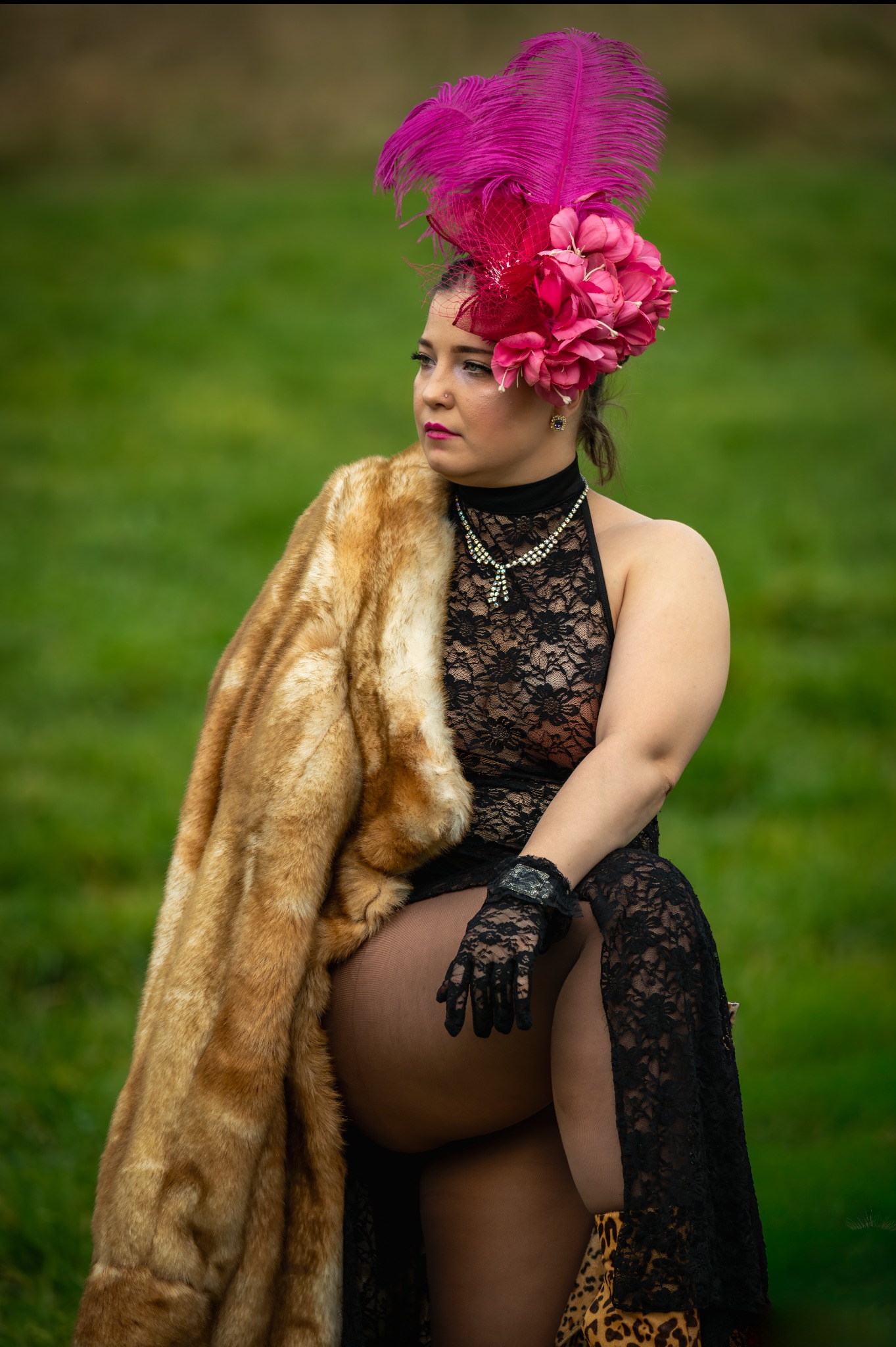 Kobieta w czarnej koronkowej sukni i futrze, z kapeluszem z piórami, siedzi na tle zieleni. Uwagę przyciąga ekstrawagancki strój i poza modelki.
