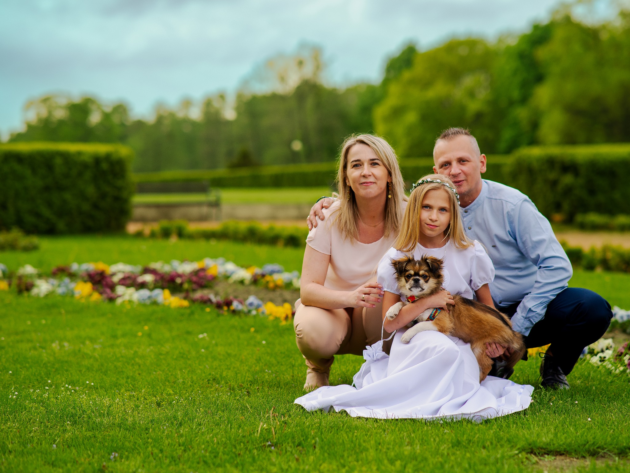 Rodzinne zdjęcie w plenerze: rodzice i córka w białej sukience trzymająca psa rasy pekińczyk na tle zieleni parku i rabat kwiatowych. Uśmiechnięte twarze.