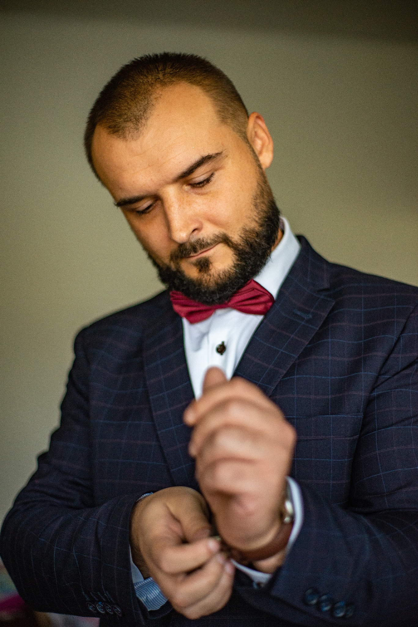 Mężczyzna w granatowej, kraciastej marynarce i bordowej muszce zapina mankiet brązowego zegarka. Ujęcie z bliska, skupione na detalu stroju.