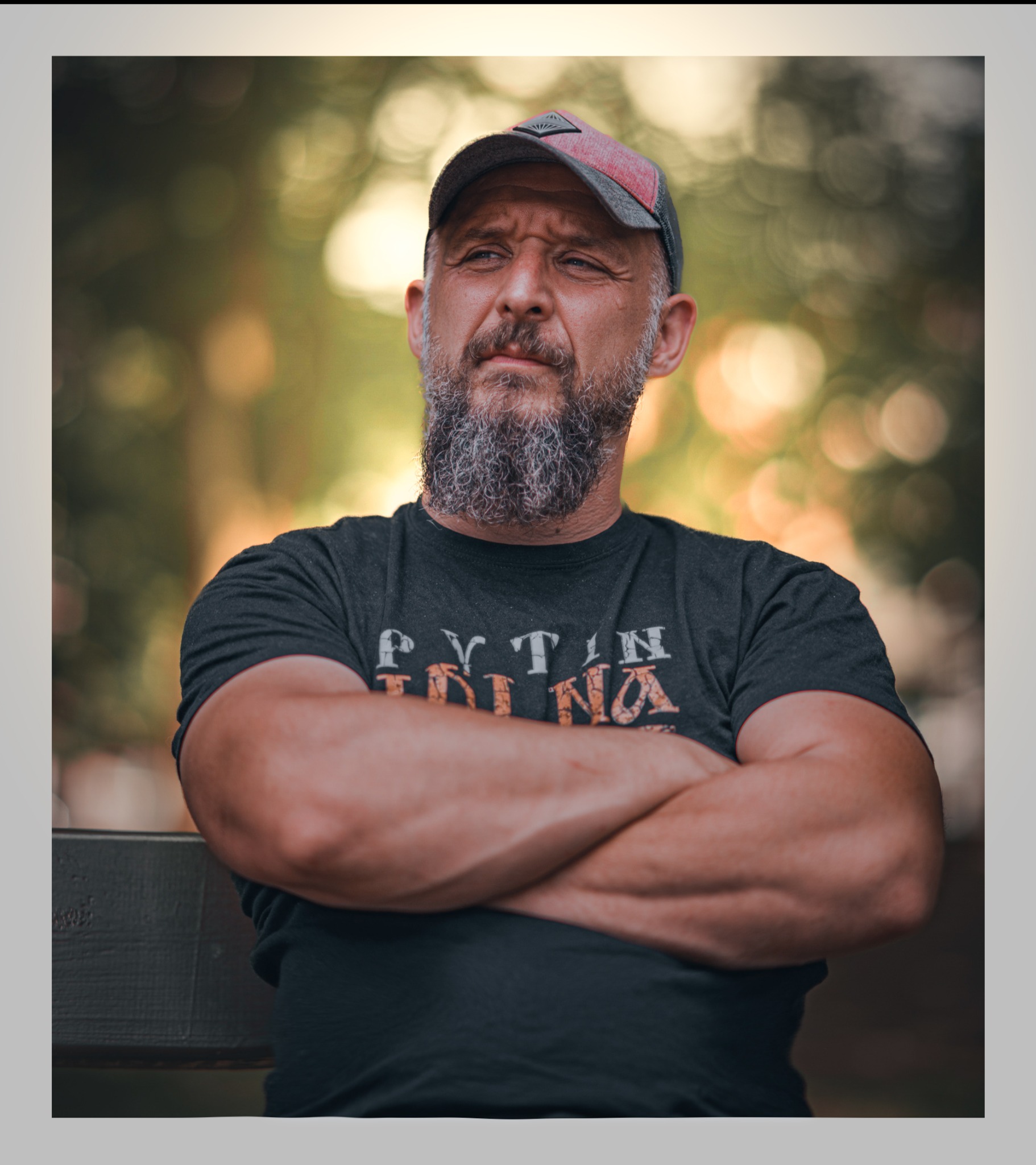 Portret brodatego mężczyzny w czarnej koszulce i czapce z daszkiem, siedzącego na ławce z założonymi rękami na tle rozmytej zieleni.