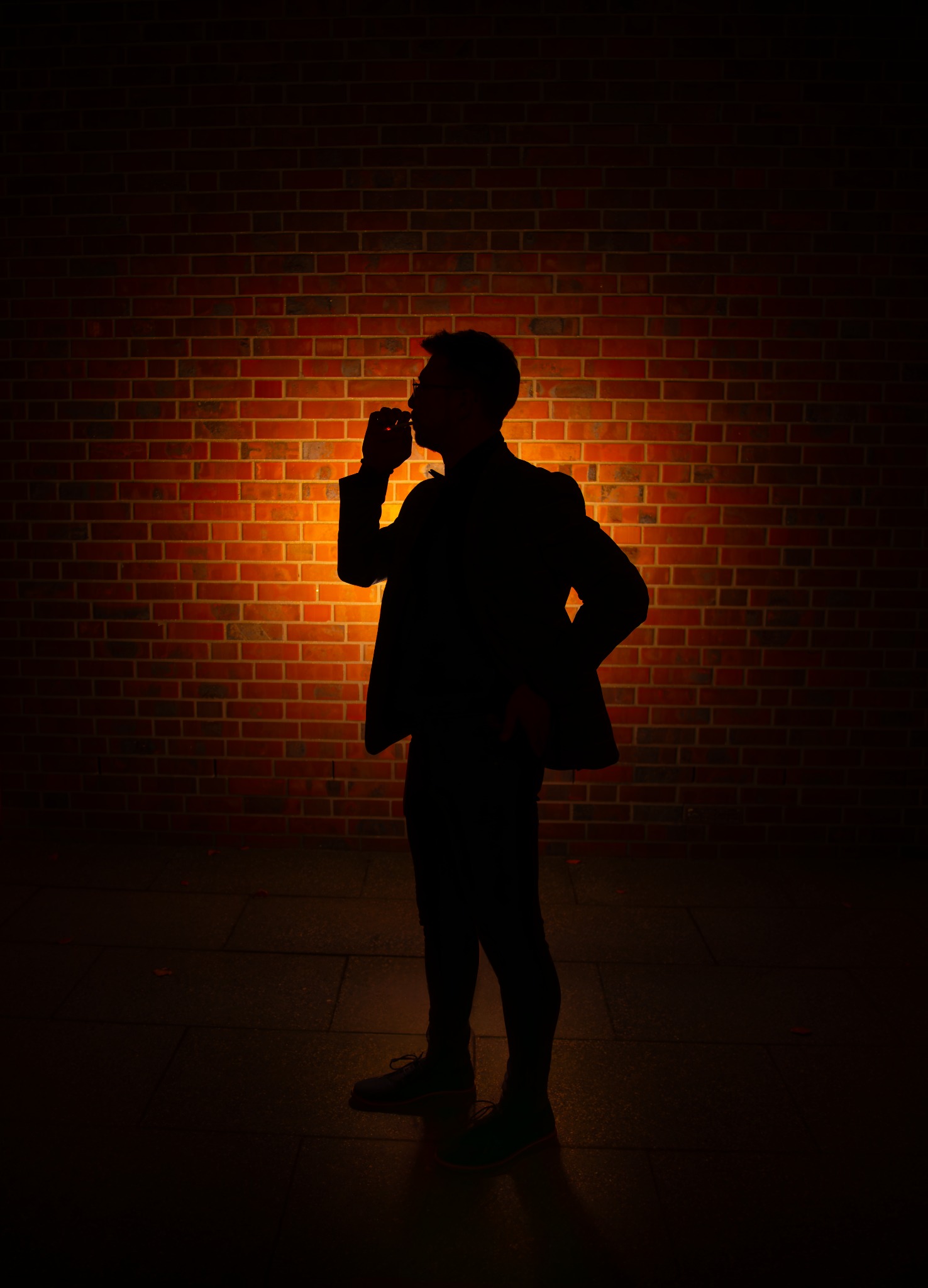 Sylwetka mężczyzny w garniturze, stojącego na tle ceglanej ściany oświetlonej ciepłym światłem, z ręką przy ustach, tworząca tajemniczy i elegancki portret.